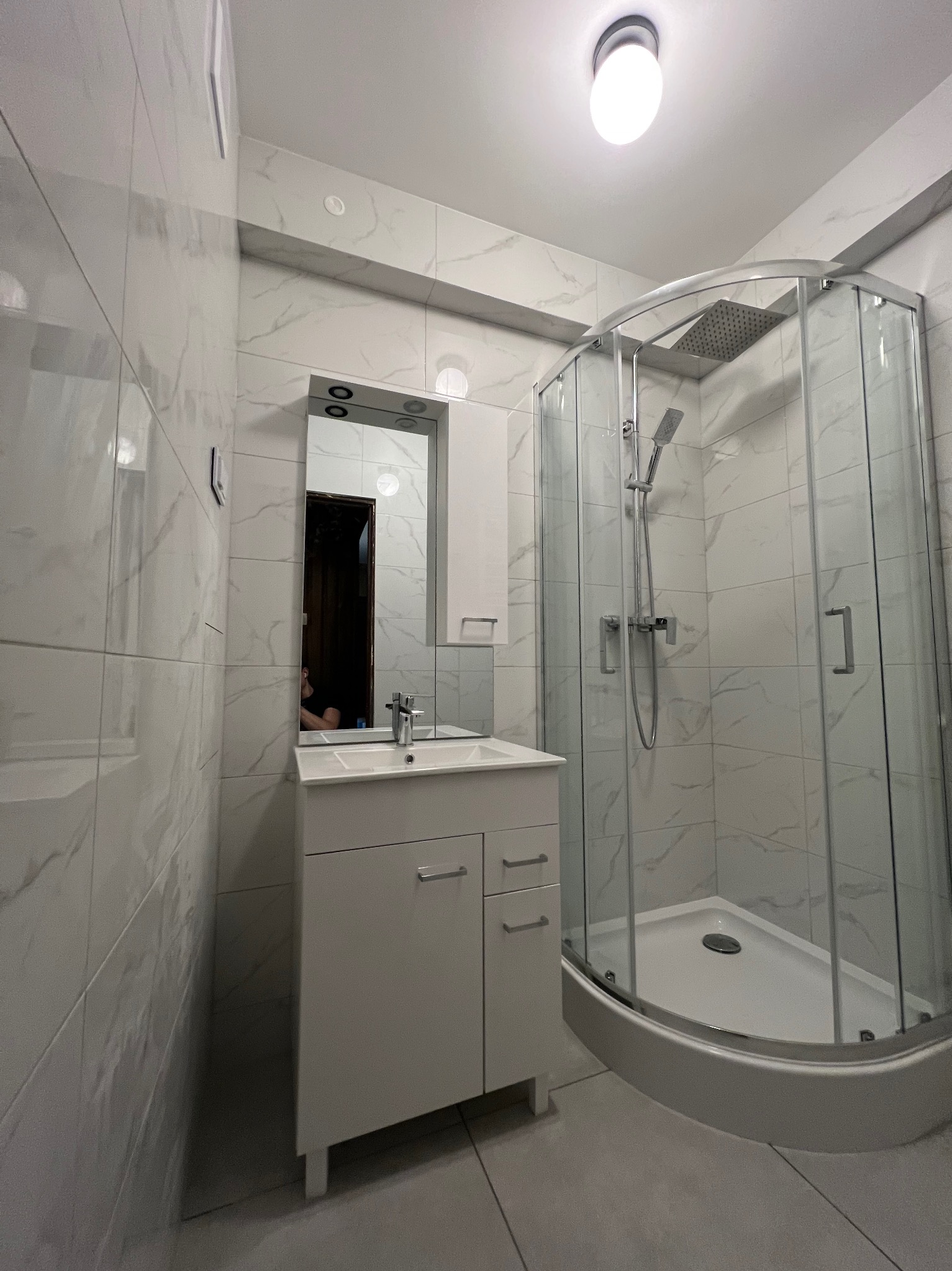 Nowoczesna łazienka po remoncie w Poznaniu: białe płytki imitujące marmur, kabina prysznicowa z deszczownicą i szafka z umywalką. Minimalistyczny design.