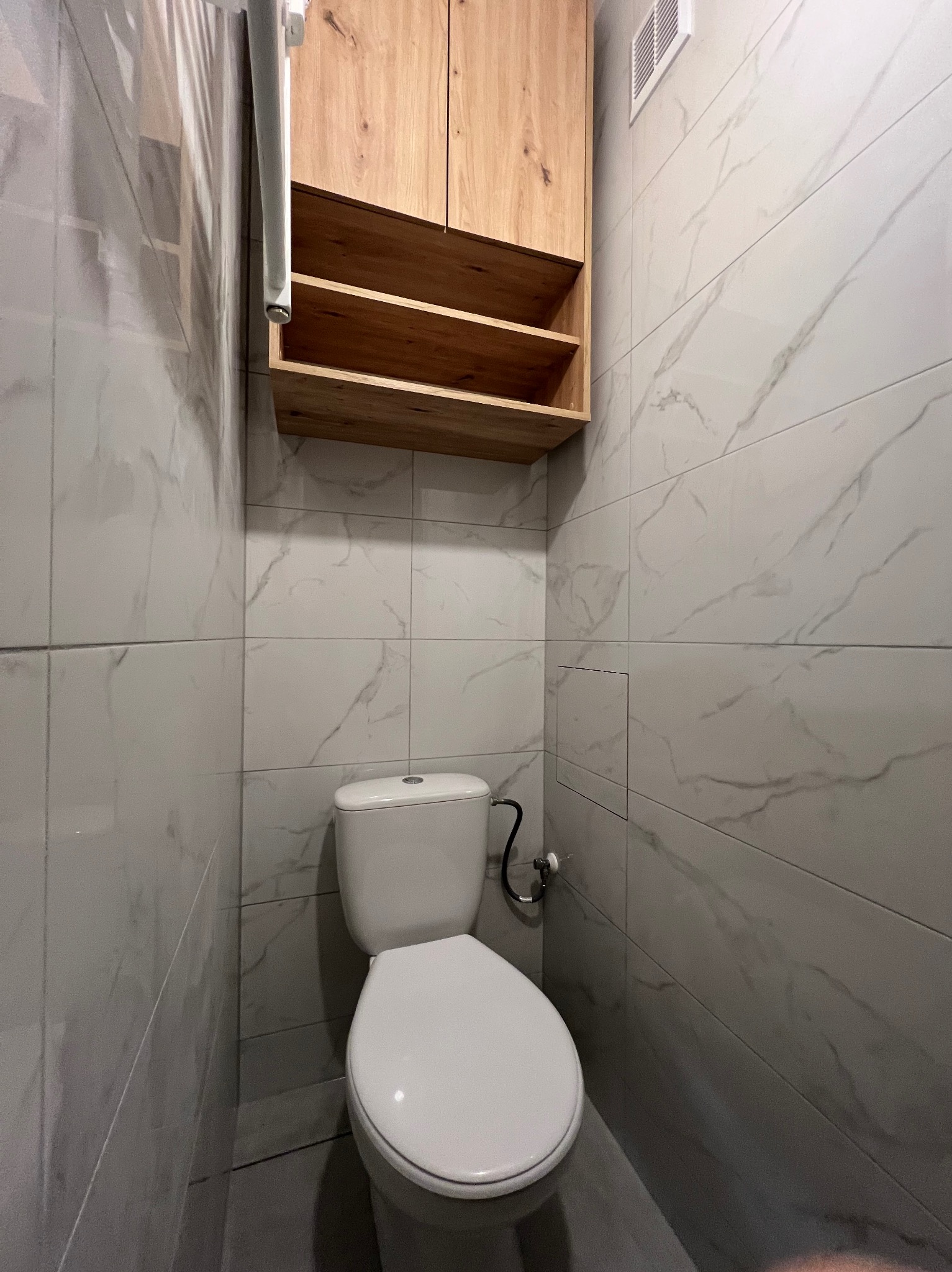 Wykończona toaleta z marmurowymi płytkami, wiszącą szafką z drewnianymi półkami i grzejnikiem drabinkowym. Minimalistyczny design, jasne wnętrze.