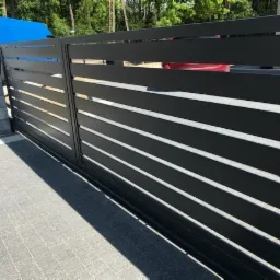 Nowoczesna, przesuwna brama wjazdowa z poziomych, czarnych profili, zamontowana na szarych, betonowych słupkach, z widoczną kostką brukową na podjeździe i zielenią w tle.