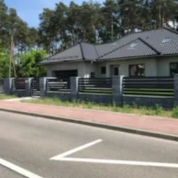 Nowoczesne ogrodzenie z poziomych, czarnych profili zamontowane na szarych, betonowych słupkach, otaczające posesję z domem jednorodzinnym. Widok z ulicy na fragment ogrodzenia i dom w tle...