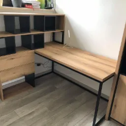 Drewniany blat biurka z czarnymi metalowymi nogami, połączony z regałem z szufladami i otwartymi półkami, całość w jasnym drewnie i czarnych akcentach, uzupełniona minimalistyczną lampką.