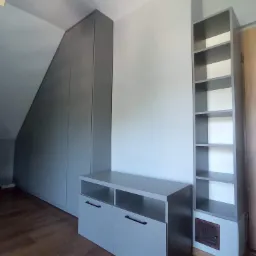 Szara zabudowa wnęki na poddaszu ze skośnym sufitem, uzupełniona szafką z czarnymi uchwytami i regałem z półkami, widoczna kratka wentylacyjna w podstawie regału.