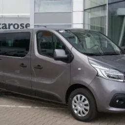 Szary Renault Trafic, wersja osobowa, zaparkowany przed salonem samochodowym z widocznym napisem 'Biuro Renault'.