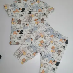 Dwu-częściowy komplet dziecięcy: koszulka z krótkim rękawem i spodnie, uszyty z białej tkaniny w pastelowe rysunki zwierząt i balonów, leżący na białym tle.