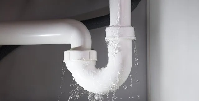 Wyciek z białego, plastikowego syfonu umywalkowego, woda kapie i rozpryskuje się, widoczne połączenia rur.
