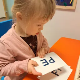 Dziewczynka w różowym swetrze siedzi przy pomarańczowym stole i przegląda karty z literami, na ścianie tablica edukacyjna z magnesami.