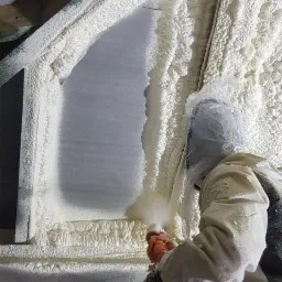 Pracownik w kombinezonie ochronnym, z pomarańczowymi rękawicami, aplikuje piankę izolacyjną na poddasze wokół okna, widoczna struktura pianki i dysza aplikatora.