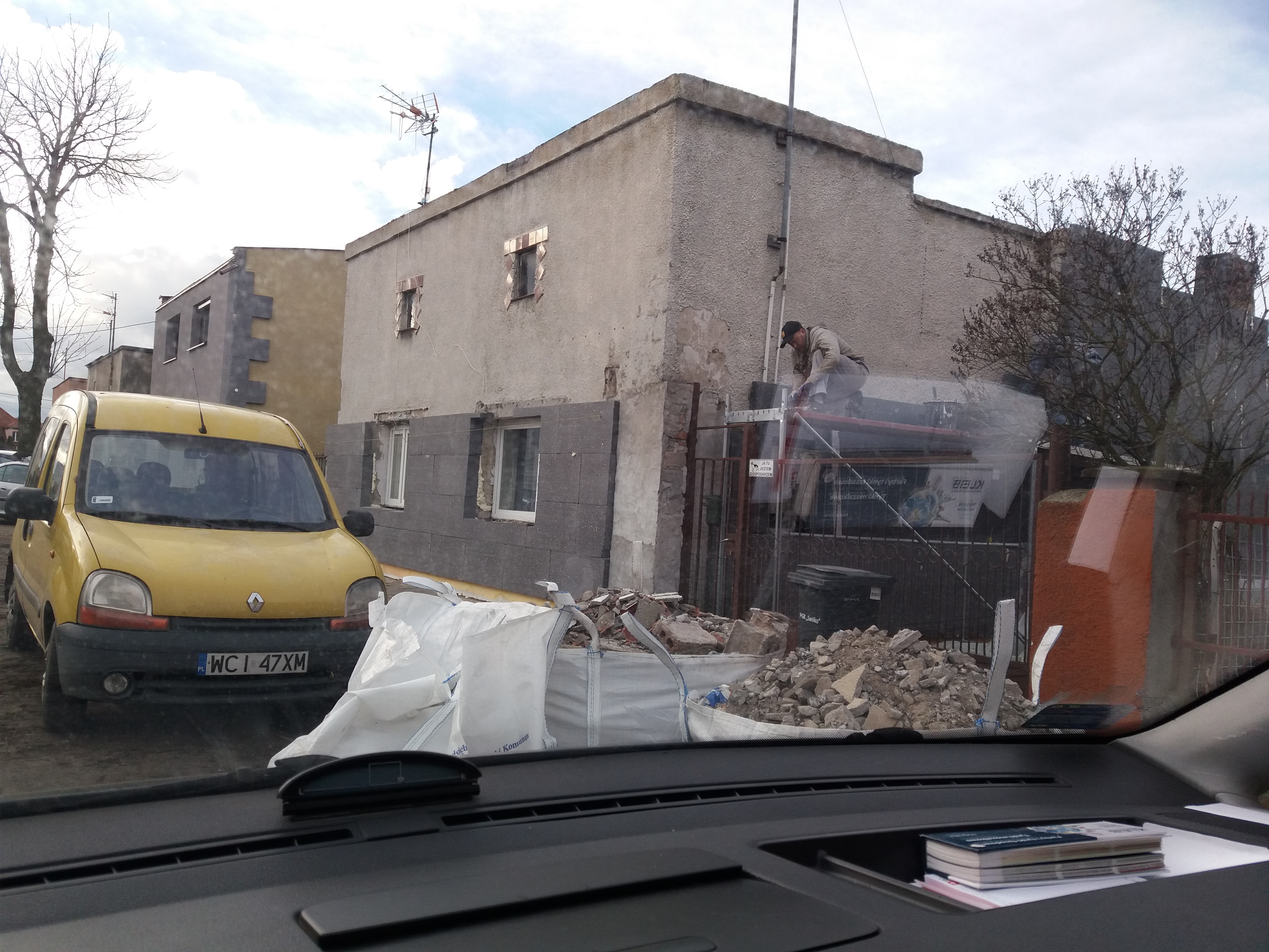 Pracownik na rusztowaniu przy ocieplanej ścianie budynku z widocznymi blokami styropianu, obok żółty samochód i worki z gruzem, ujęcie z wnętrza pojazdu.