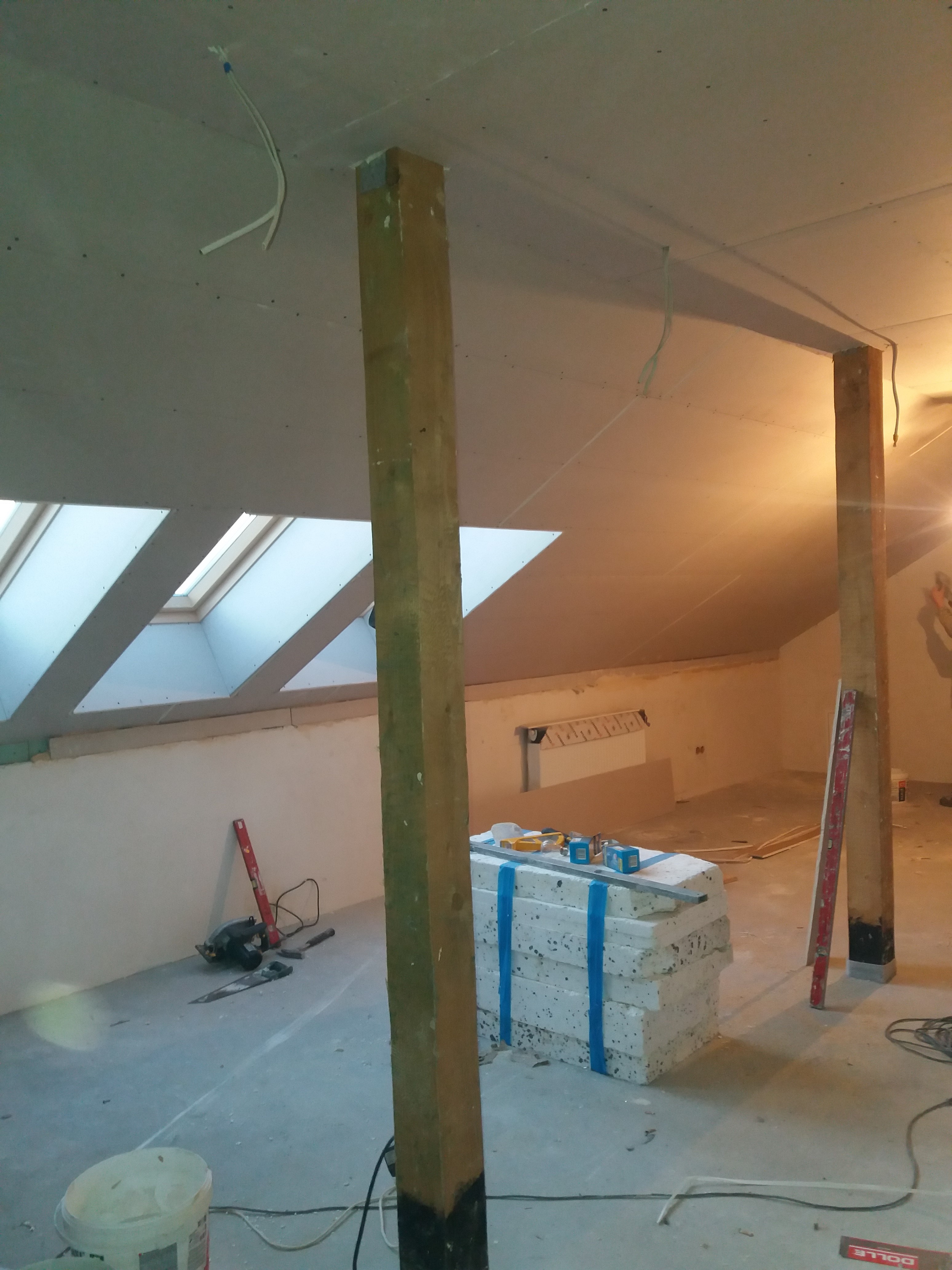 Poddasze w trakcie prac wykończeniowych, widoczne drewniane słupy wzmacniające konstrukcję, okna dachowe, przygotowane płyty styropianowe, narzędzia i poziomice, surowe ściany i sufity z płyt...