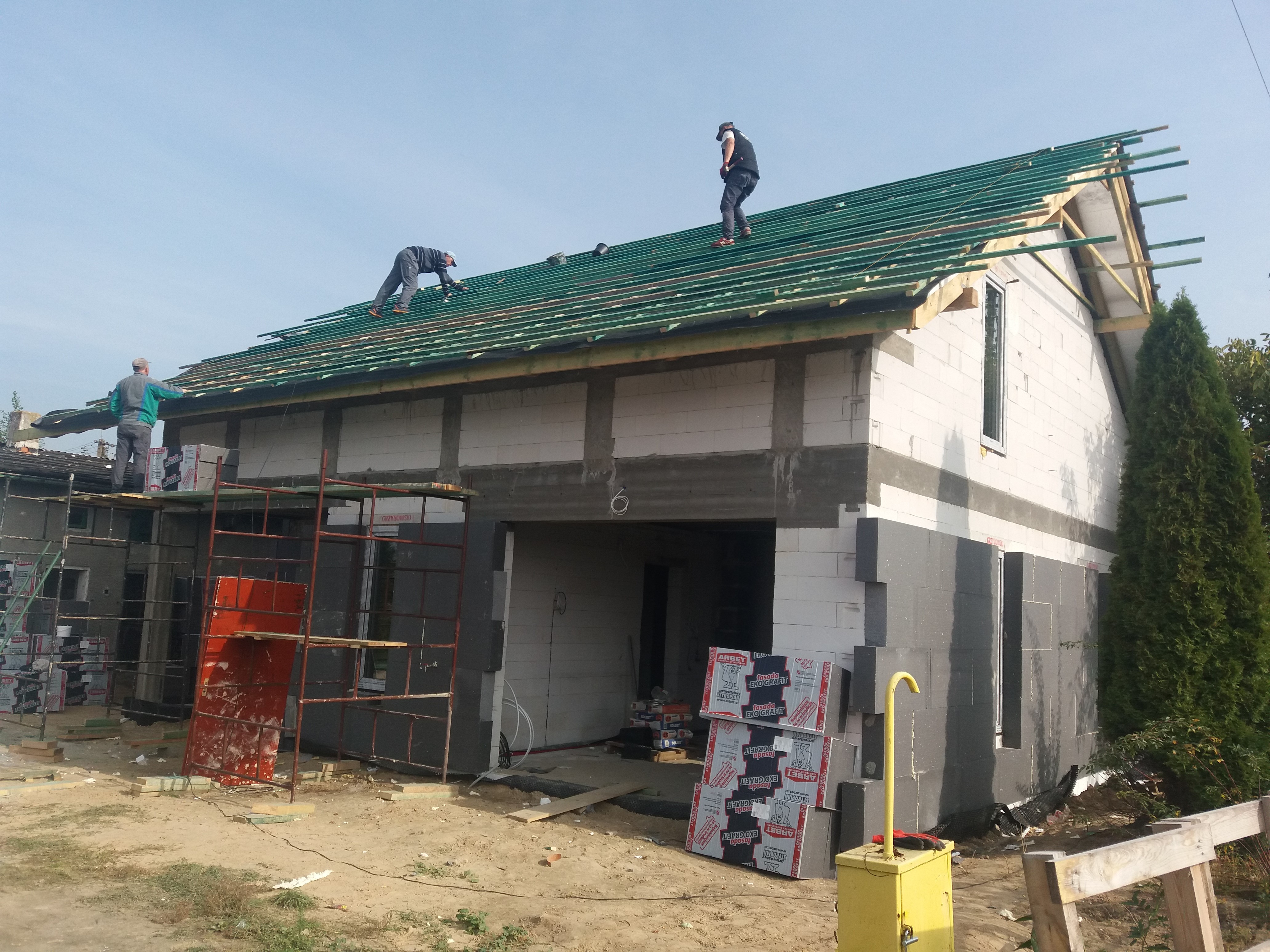 Budowa domu jednorodzinnego w trakcie: ekipa montuje konstrukcję dachu z zieloną membraną, ściany z białego bloczka częściowo ocieplone szarym styropianem, na ziemi paczki styropianu w opakowaniach...