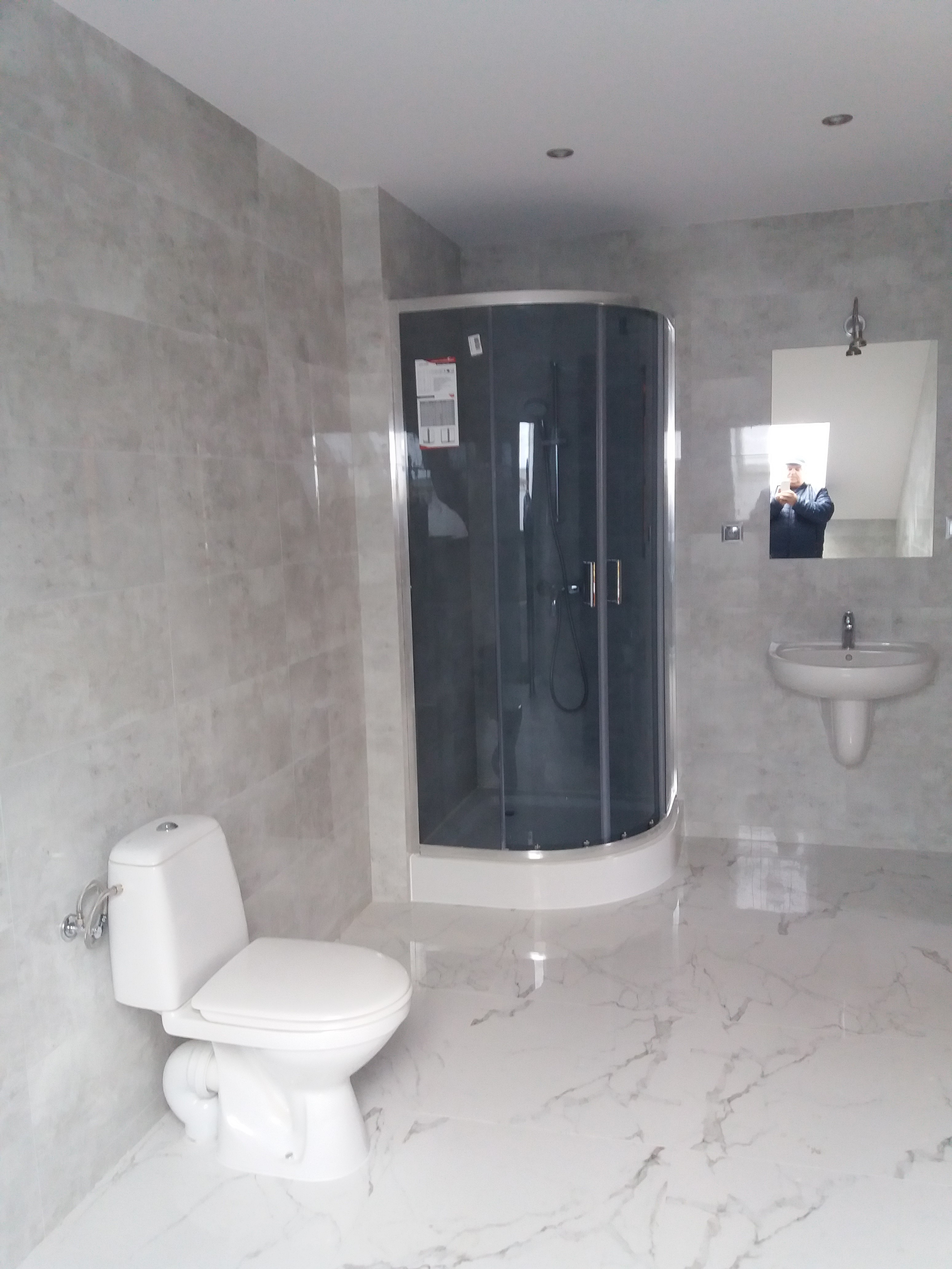 Wykończona łazienka w odcieniach szarości i bieli z kabiną prysznicową narożną, umywalką i toaletą, widok na fragment osoby robiącej zdjęcie w lustrze.
