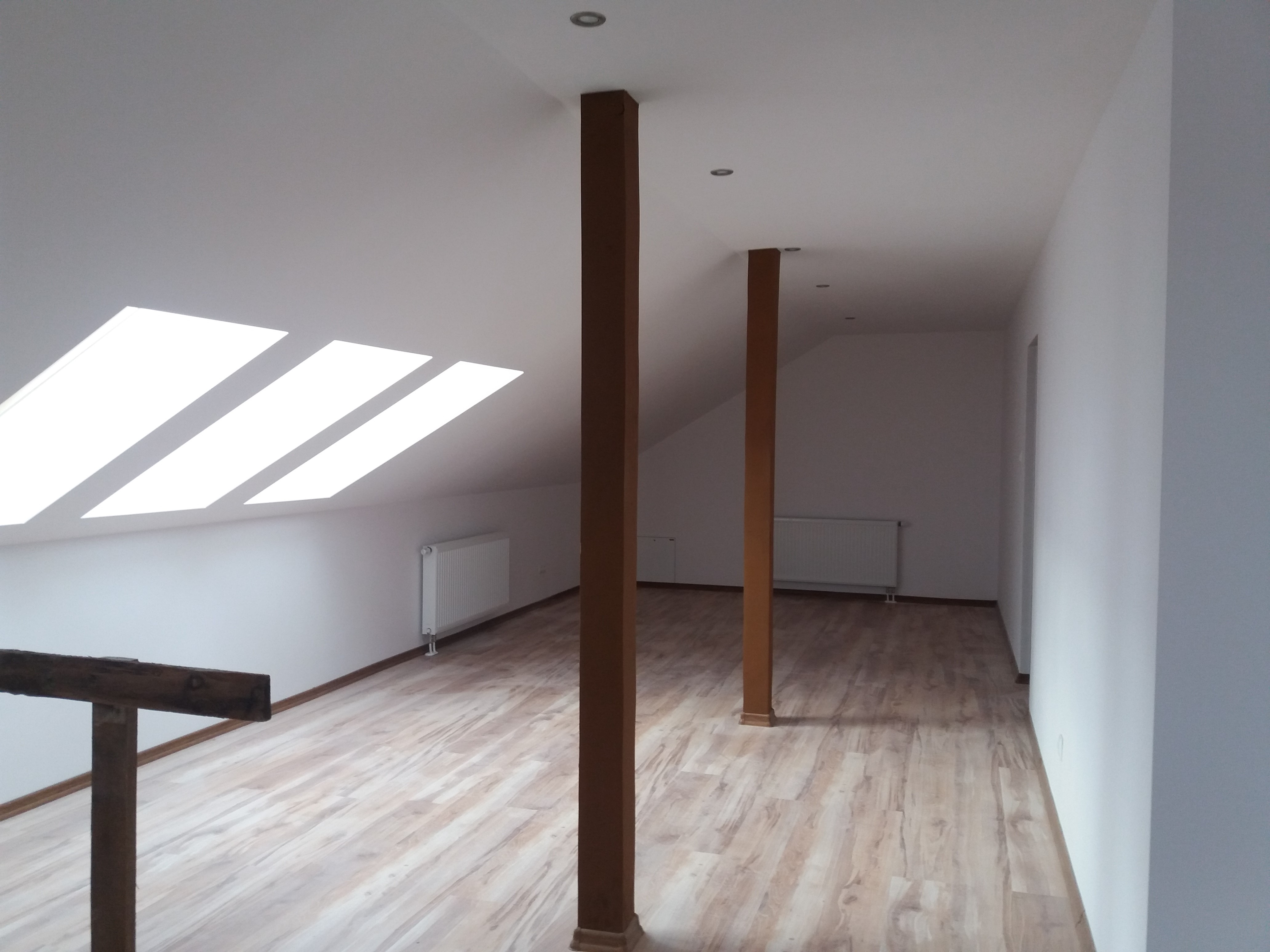 Wykończone poddasze z drewnianą podłogą, pomalowanymi na biało ścianami, drewnianymi słupami i oknami dachowymi w skosach.