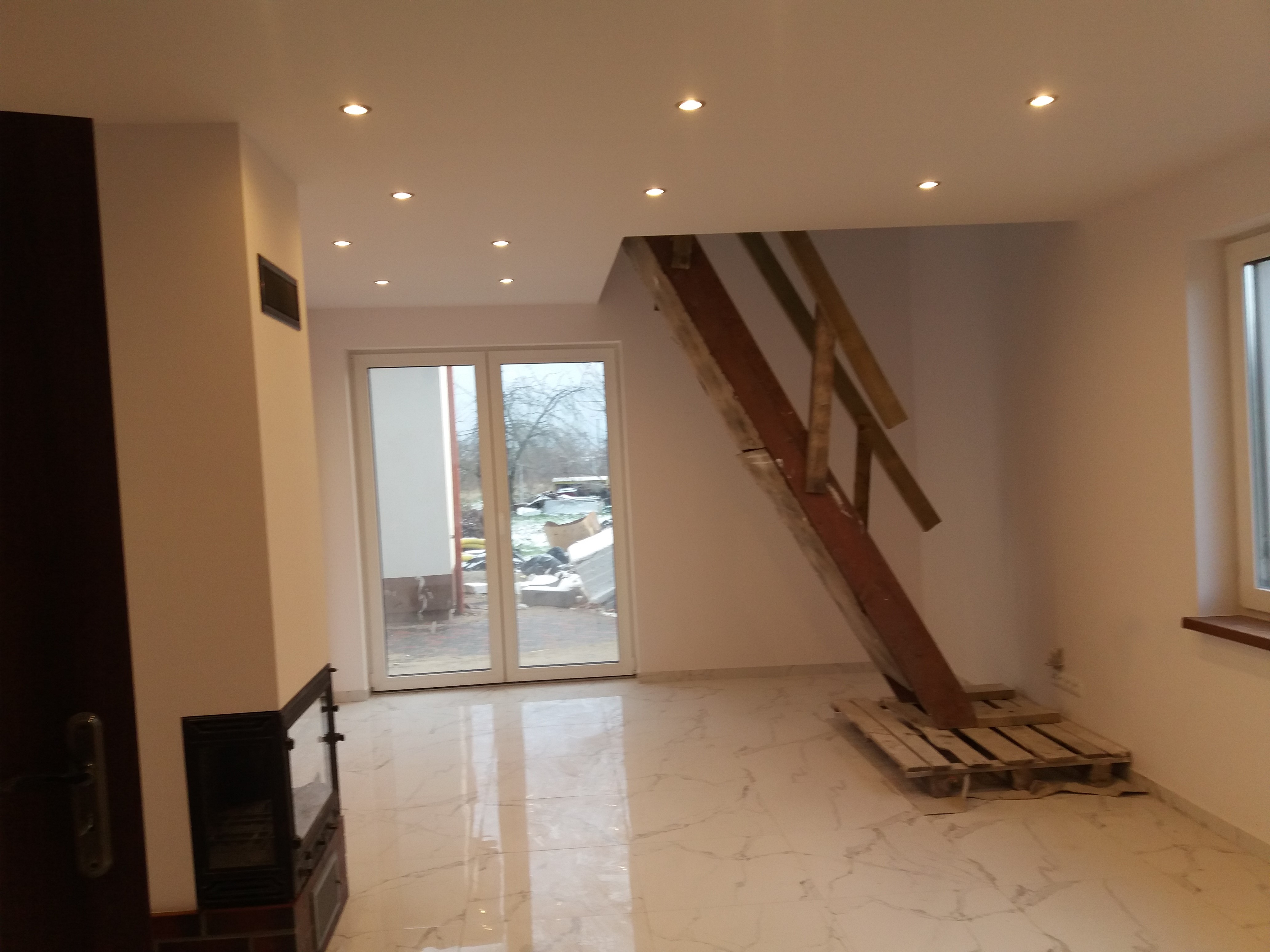 Wnętrze w trakcie remontu we Włocławku: białe ściany, marmurowe płytki podłogowe, prowizoryczne drewniane schody oparte na palecie i widok na podwórko przez rozsuwane drzwi balkonowe.