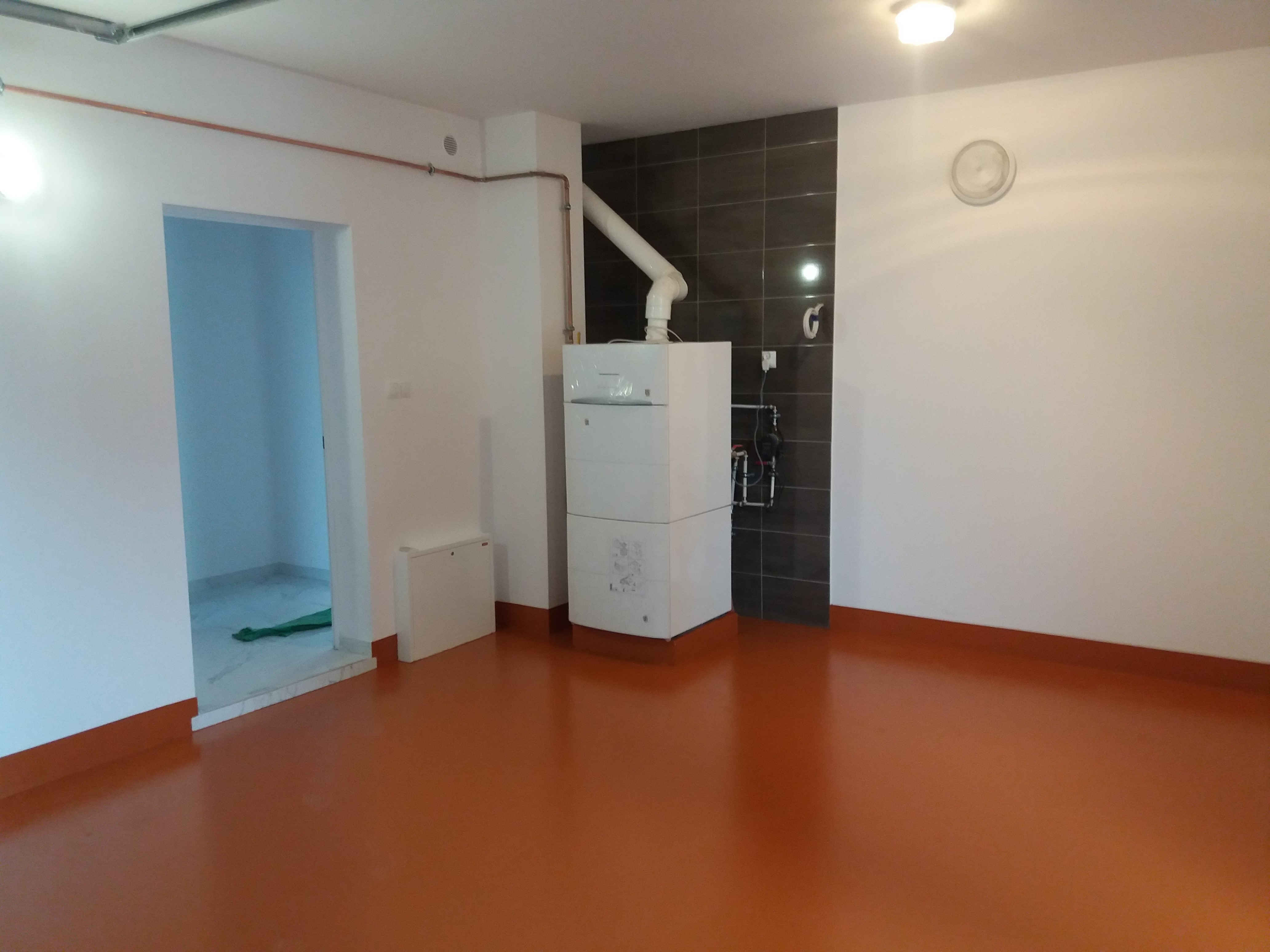 Pomieszczenie po remoncie z nową pomarańczową podłogą żywiczną, piecem grzewczym i fragmentem wykończonej ściany z ciemnymi płytkami. Widoczne rury grzewcze na ścianie i wejście do innego...