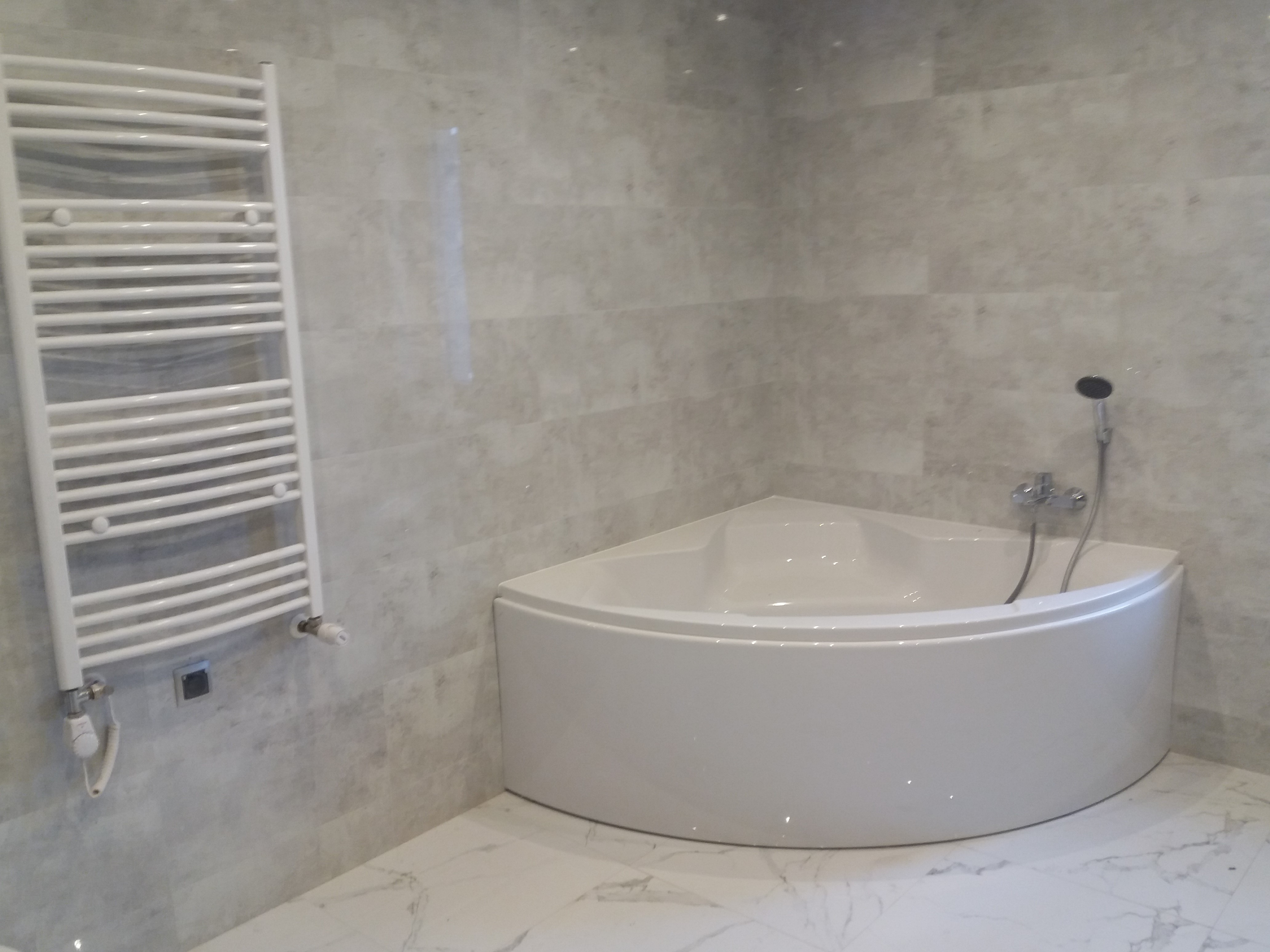 Narożna wanna z baterią i słuchawką prysznicową, biały grzejnik drabinkowy na ścianie z szarych płytek imitujących marmur, marmurowa podłoga.