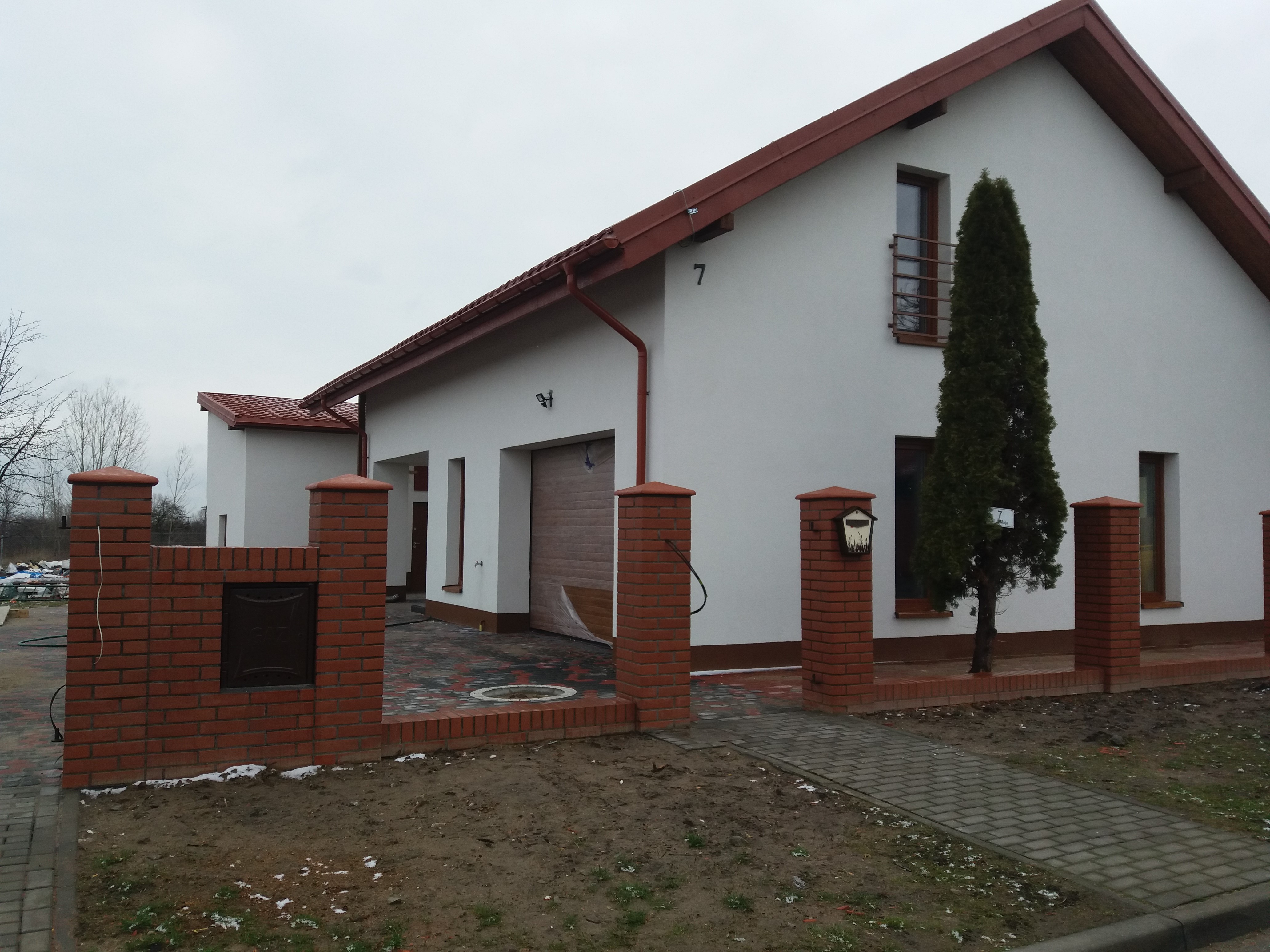 Biały dom jednorodzinny z czerwoną dachówką, ceglanym ogrodzeniem, numerem 7 na elewacji i małą, zieloną tują przed oknem.