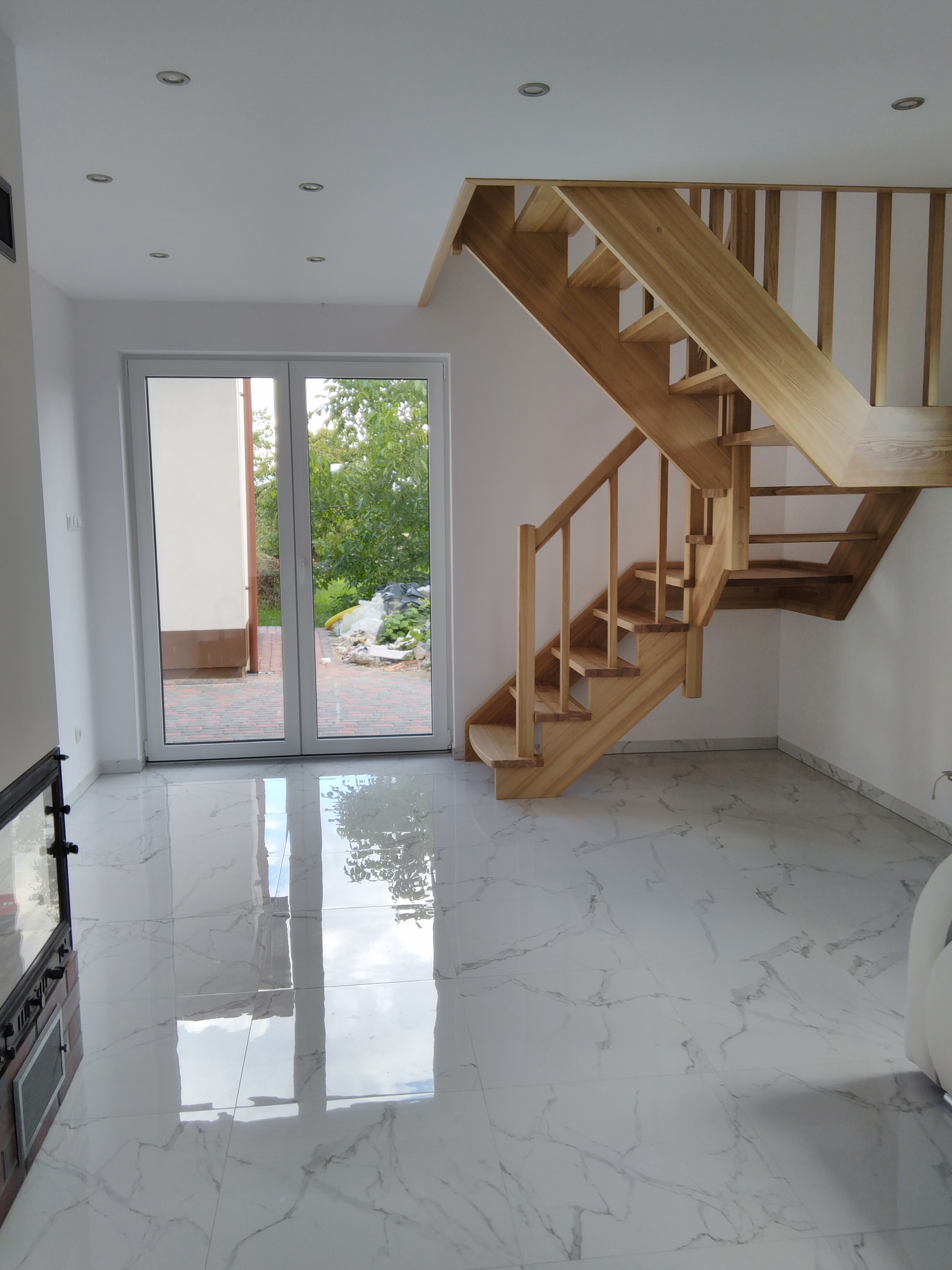 Jasne wnętrze z marmurową podłogą, drewnianymi schodami i dużym oknem tarasowym wychodzącym na ogród.
