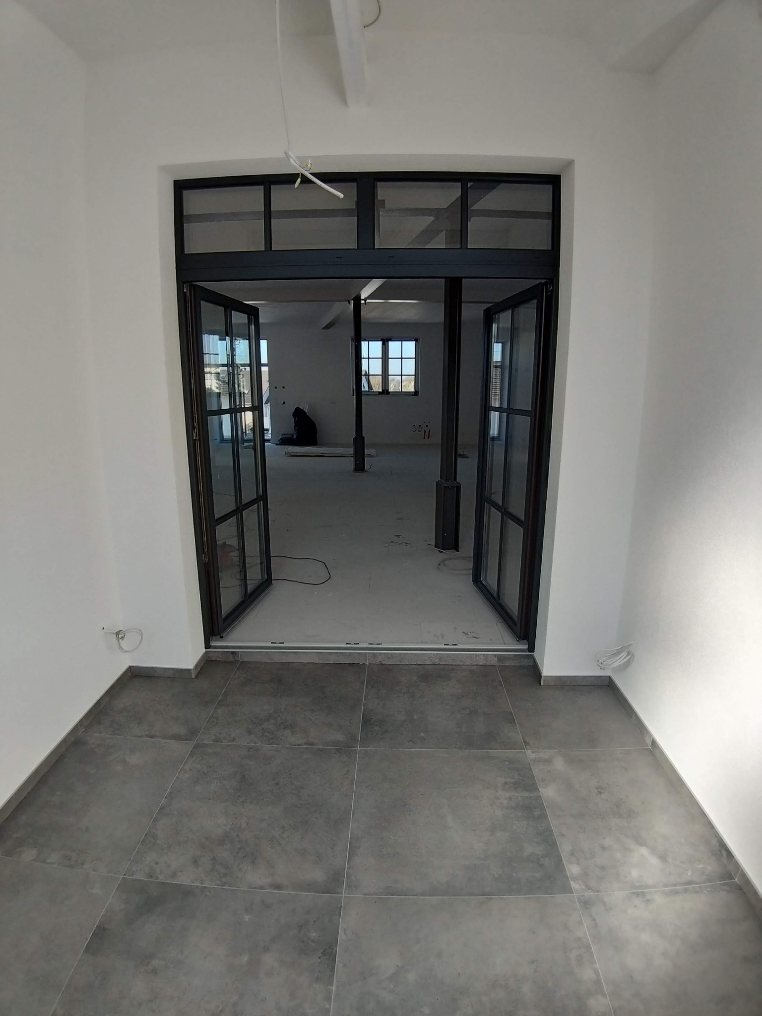 Wnętrze w trakcie odświeżania: widok z korytarza na otwarte, przeszklone drzwi prowadzące do pomieszczenia z białymi ścianami i szarą podłogą. W głębi widoczna osoba w ciemnym ubraniu...
