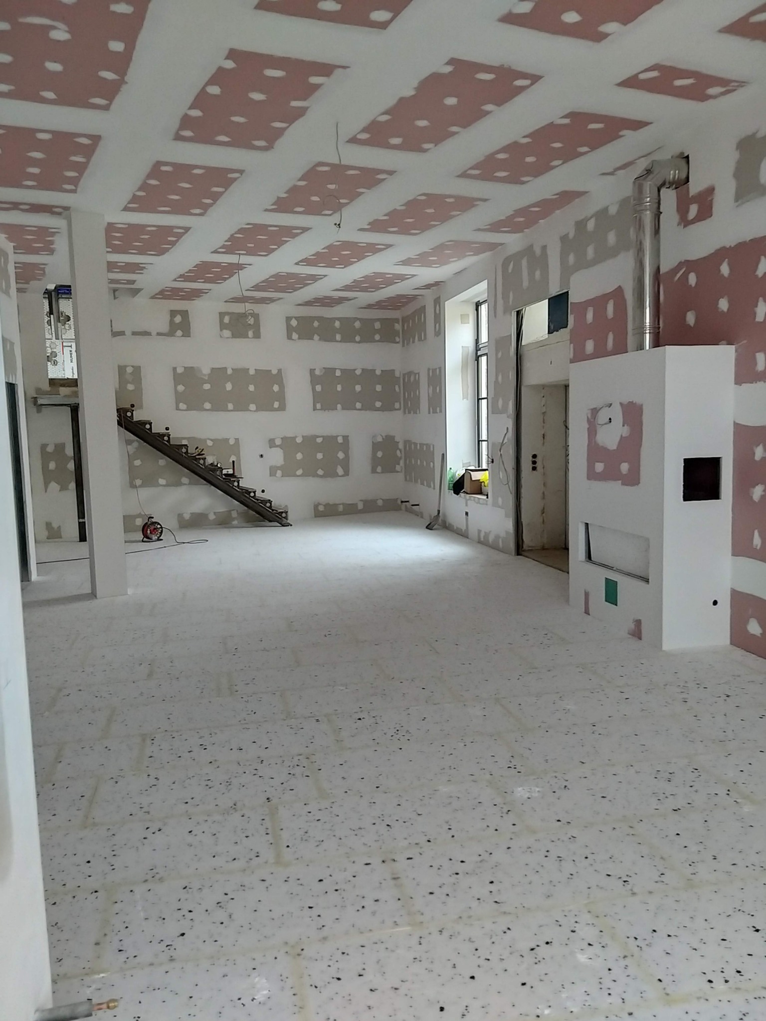 Wnętrze w trakcie prac malarskich i wykończeniowych: ściany i sufit z widocznymi fragmentami płyt gipsowo-kartonowych, przygotowane do malowania, surowa stalowa konstrukcja schodów, biały kominek...