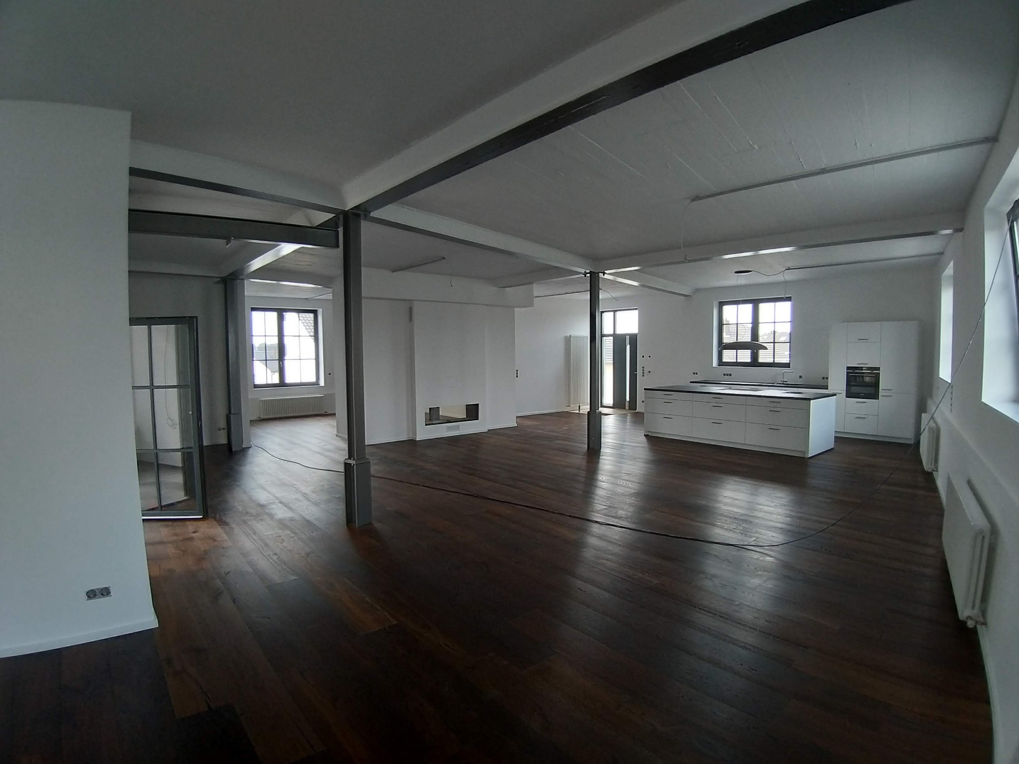 Przestronne, jasne wnętrze loftu z drewnianą podłogą, białymi ścianami i metalowymi belkami konstrukcyjnymi, częściowo wyposażone w białe meble kuchenne i grzejniki.