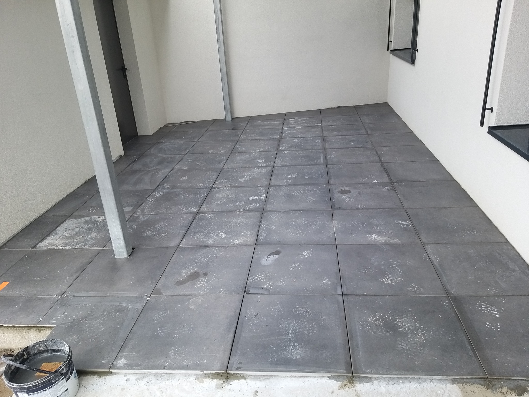 Układanie szarych płytek na tarasie, widoczne ślady butów na powierzchni płytek, wiadro z zaprawą w rogu, metalowy słup wbudowany w posadzkę.