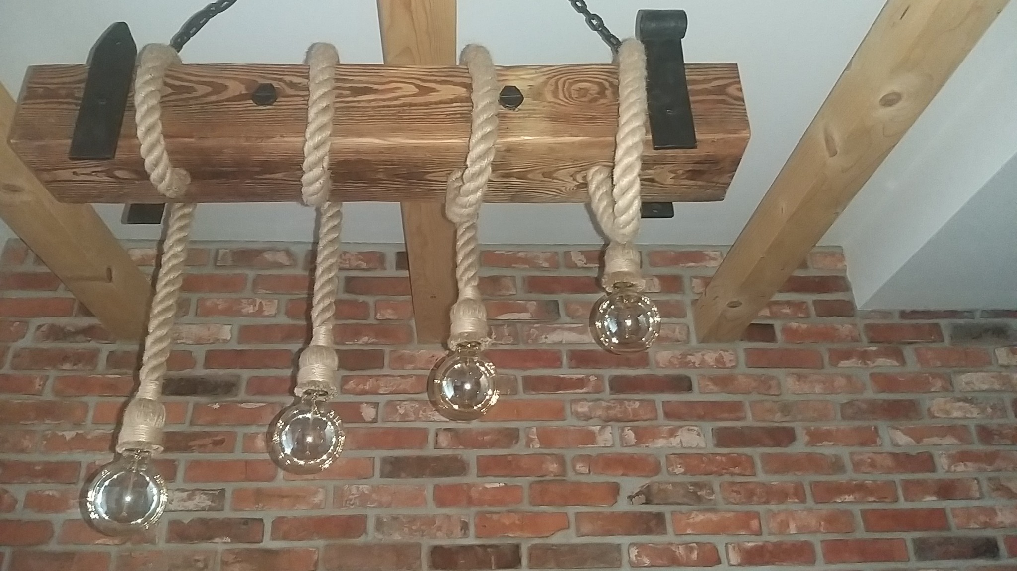 Rustykalna lampa wisząca z drewnianej belki i grubych lin z żarówkami Edisona na tle ceglanej ściany.