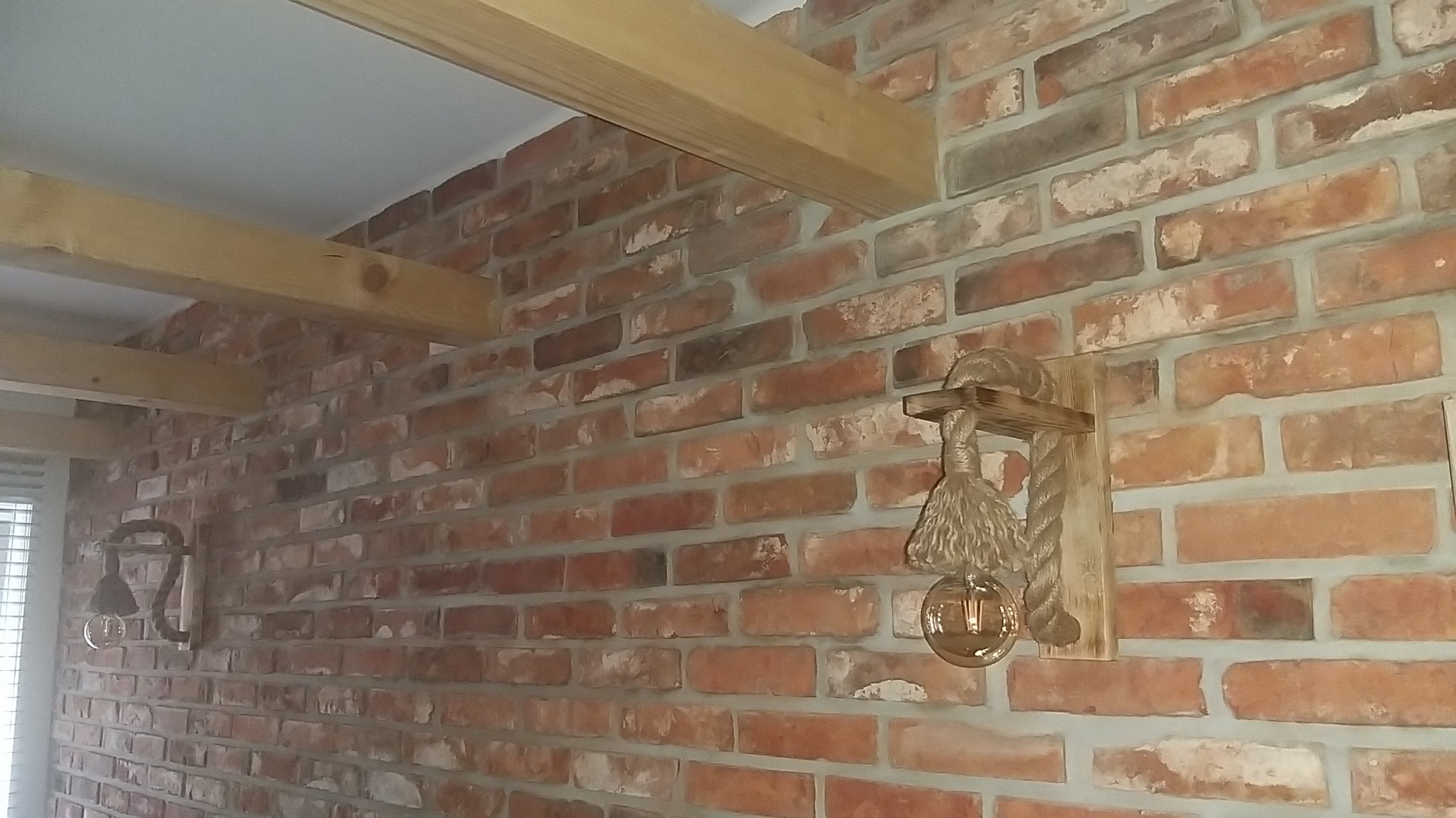 Ściana z czerwonej cegły z drewnianymi belkami stropowymi i dekoracyjnym oświetleniem ściennym w stylu marynistycznym z liną jutową.