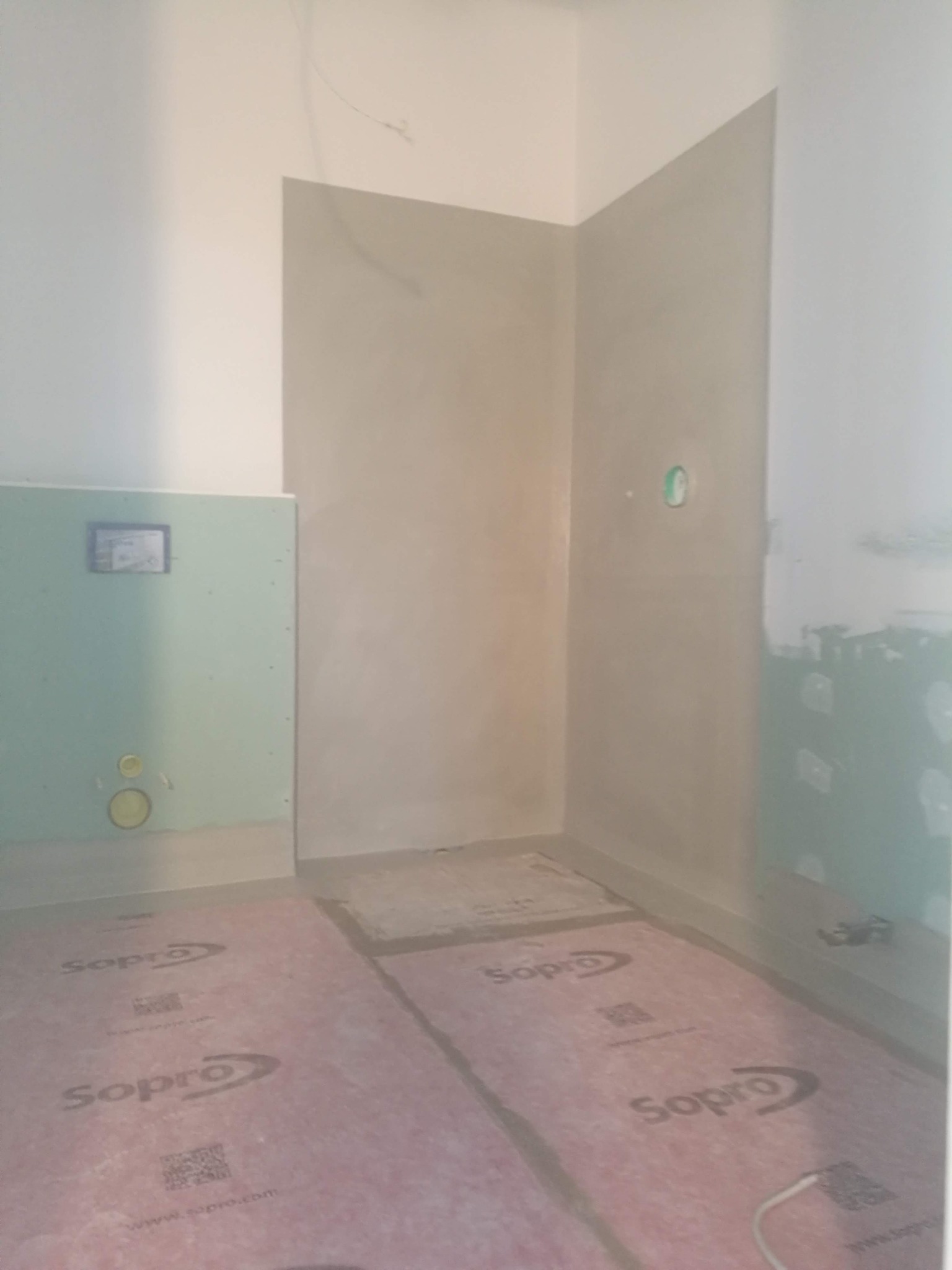 Przygotowanie ścian w łazience do położenia płytek, widoczne fragmenty ścian z nałożonym tynkiem i płyty gipsowo-kartonowe, podłoga zabezpieczona folią ochronną marki Sopro.