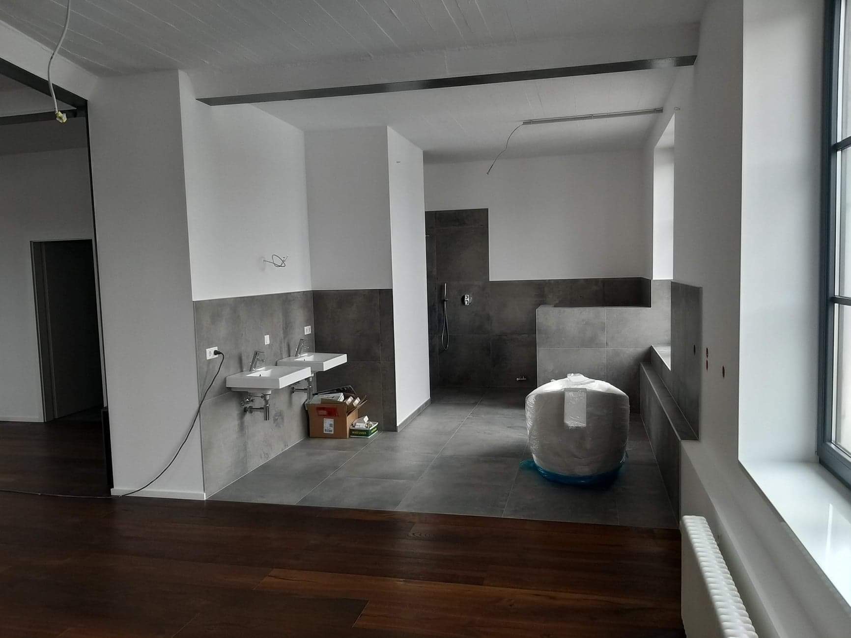 Wnętrze łazienki w trakcie remontu, z widocznymi dwoma białymi umywalkami, szarą glazurą do połowy ściany, zapakowanym bojlerem i drewnianą podłogą w odcieniach brązu.