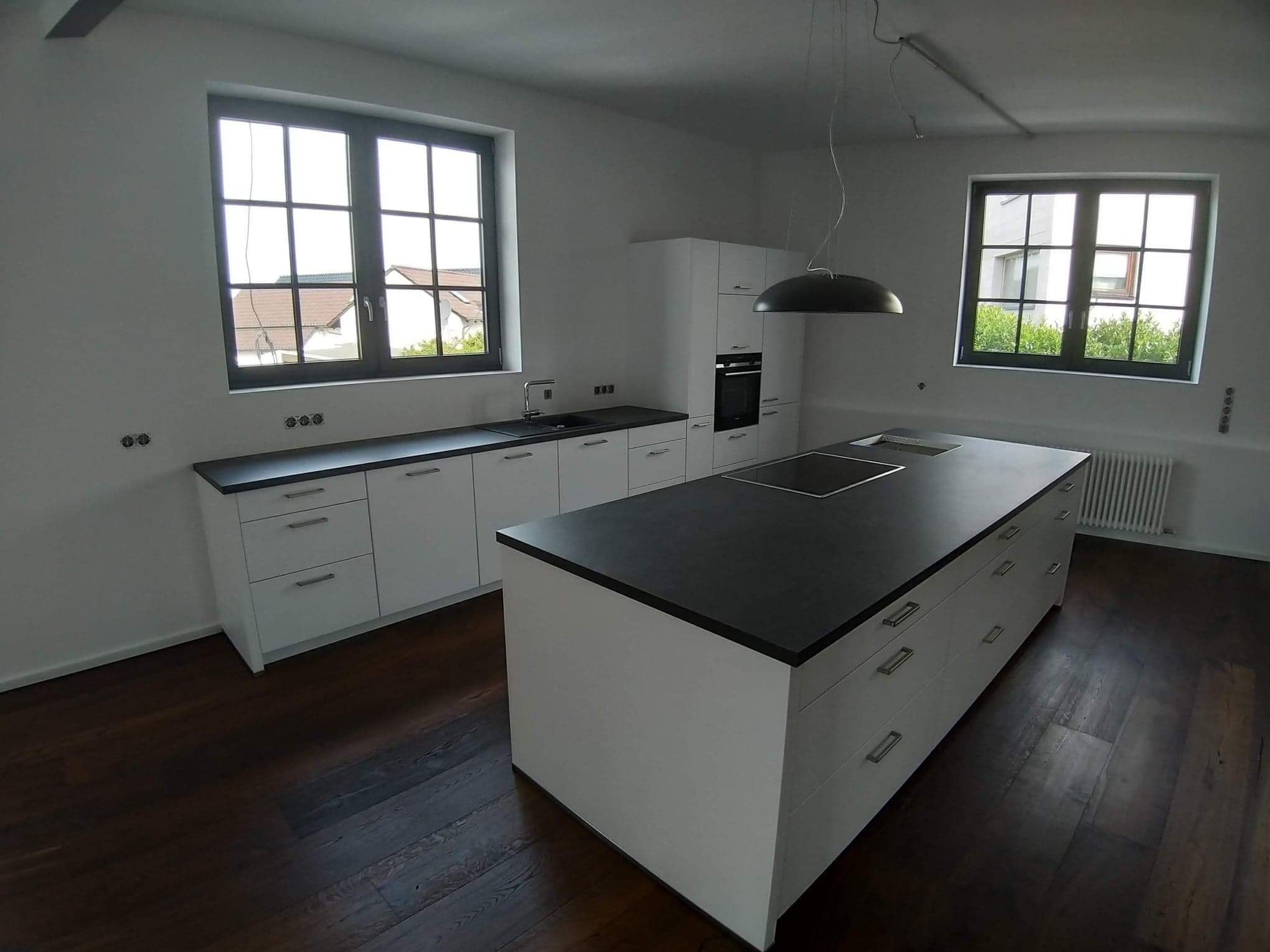 Nowoczesna kuchnia z białymi szafkami i czarnym blatem, wyspa kuchenna na ciemnej drewnianej podłodze, duże okna z widokiem na zewnątrz.