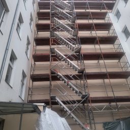 Sebastian Napierała - Elewacja budynku w trakcie prac, z widocznymi rusztowaniami i schodami, częściowo osłonięta folią ochronną.