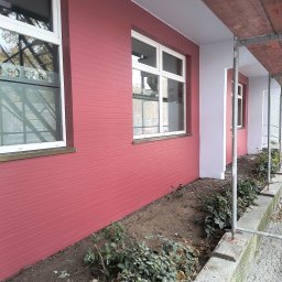 Sebastian Napierała - Świeżo pomalowana elewacja budynku w kolorze ceglastym, z białymi ramami okiennymi, widoczne rusztowania i fragment rabaty z zielenią.