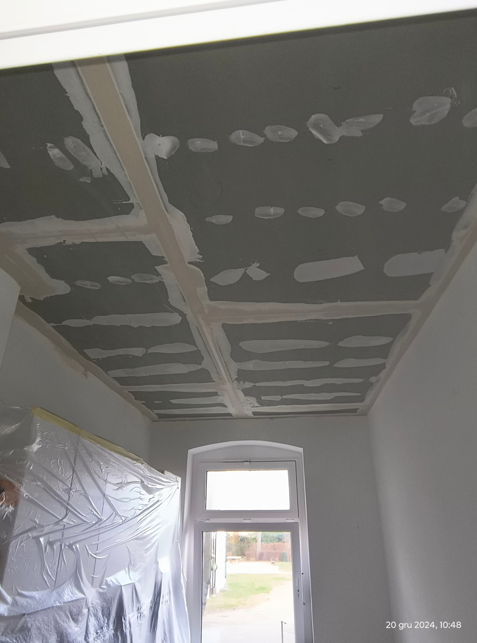 Sufit z płyt kartonowo-gipsowych w trakcie montażu, widoczne szpachlowane łączenia, zabezpieczone folią elementy pomieszczenia oraz okno.