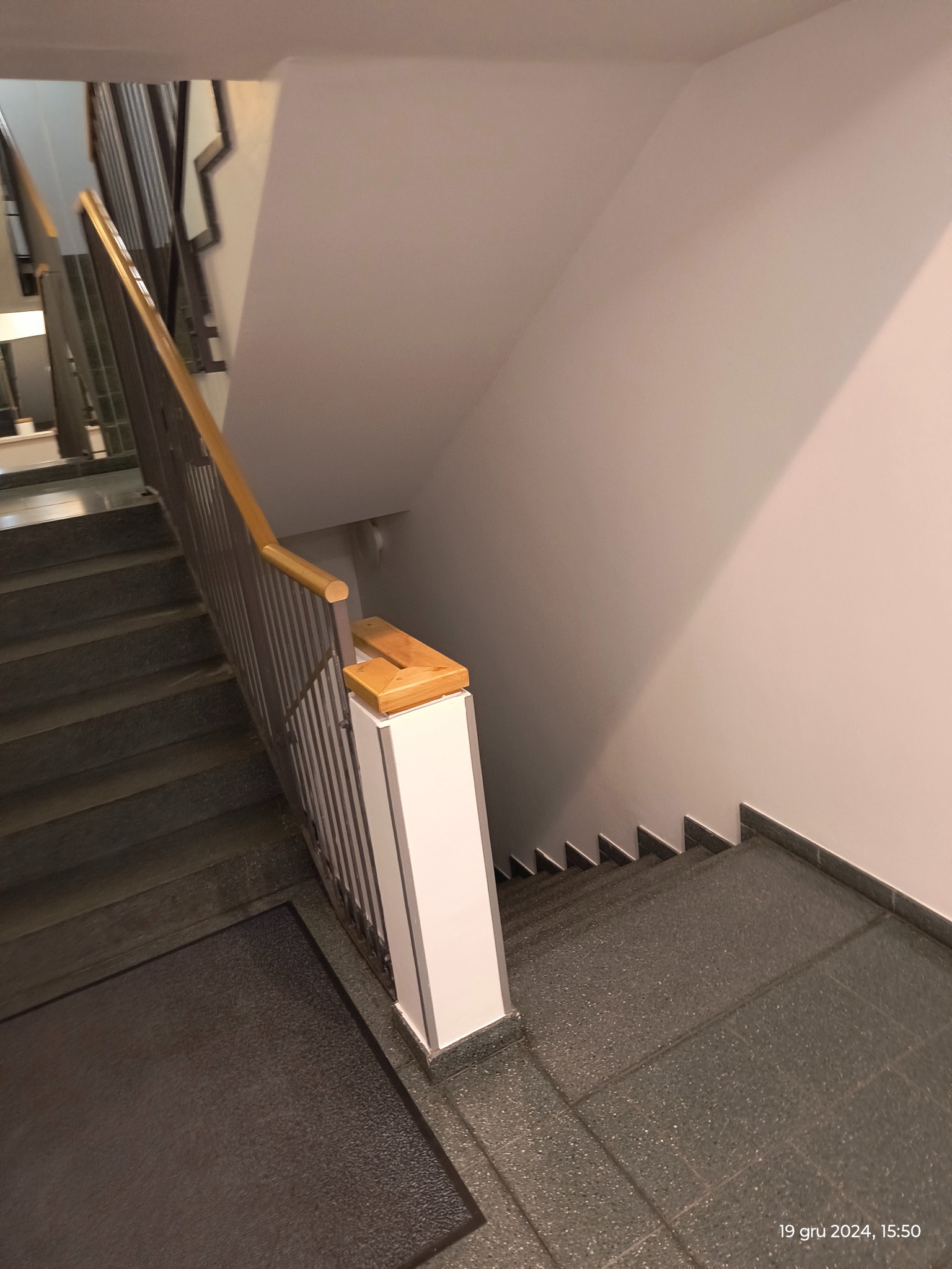 Klatka schodowa z metalową balustradą i drewnianym poręczą, widok z góry na schody pokryte szarą terakotą.