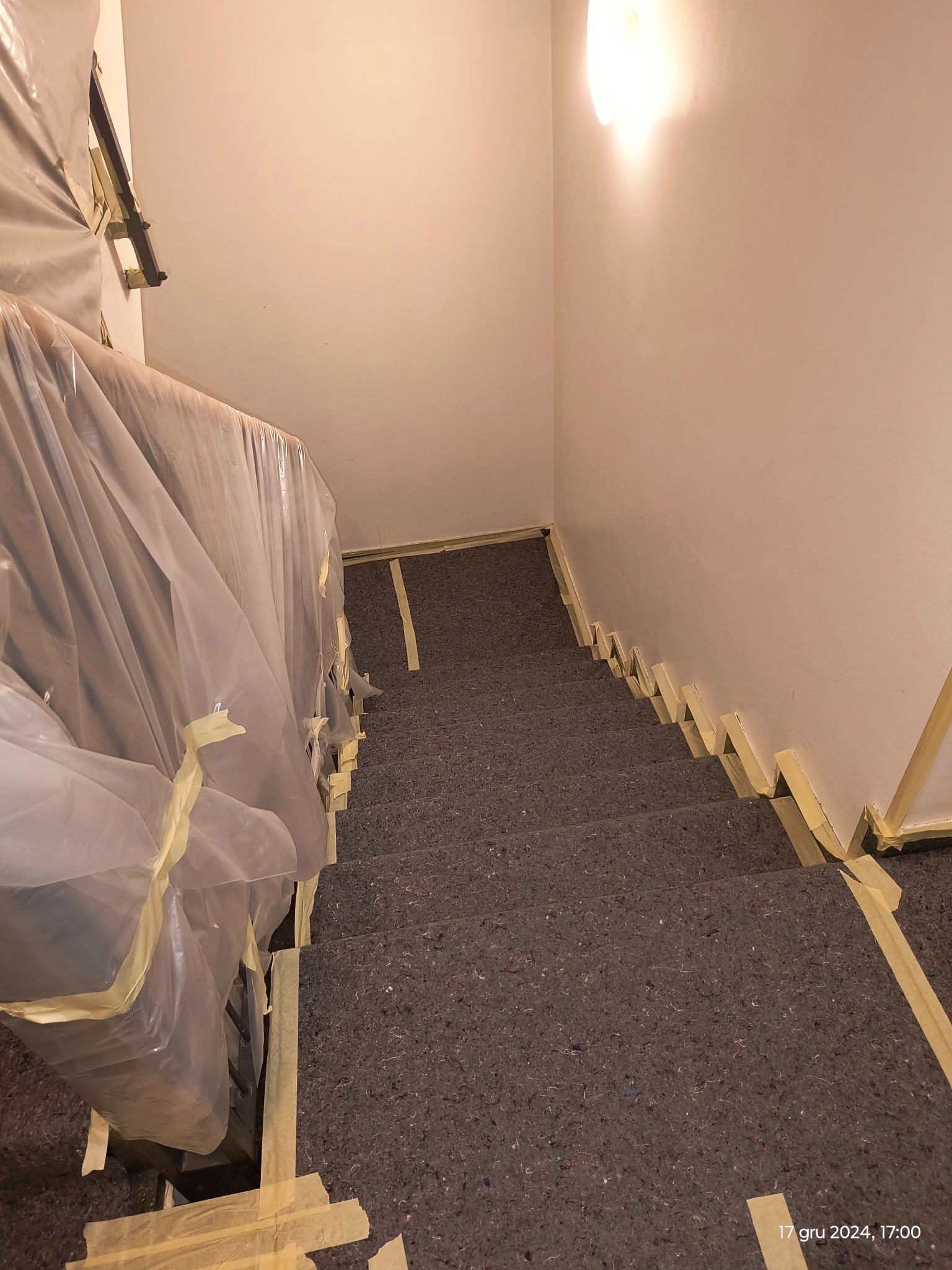 Klatka schodowa zabezpieczona folią i taśmą malarską przed pracami wykończeniowymi, widok z góry na schody pokryte materiałem ochronnym.