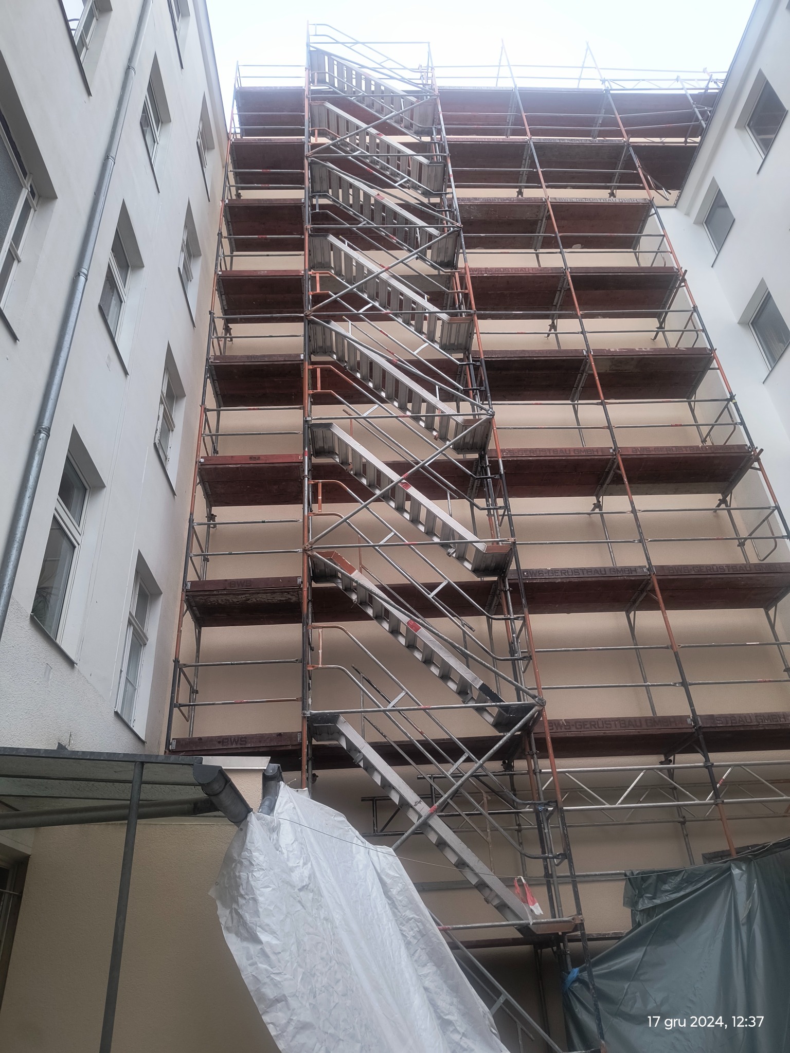 Elewacja budynku w trakcie prac, z widocznymi rusztowaniami i schodami, częściowo osłonięta folią ochronną.