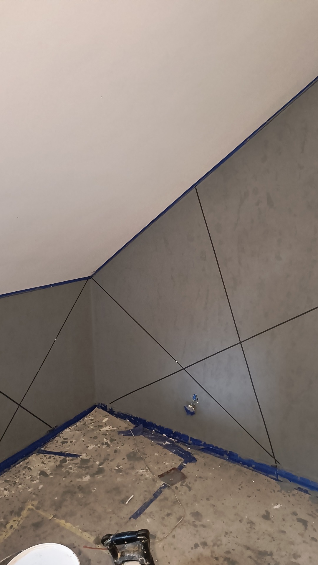 Ściana w szarym odcieniu z geometrycznym wzorem z czarnych linii, zabezpieczona niebieską taśmą malarską, widoczne ślady prac na podłodze i narzędzia malarskie.