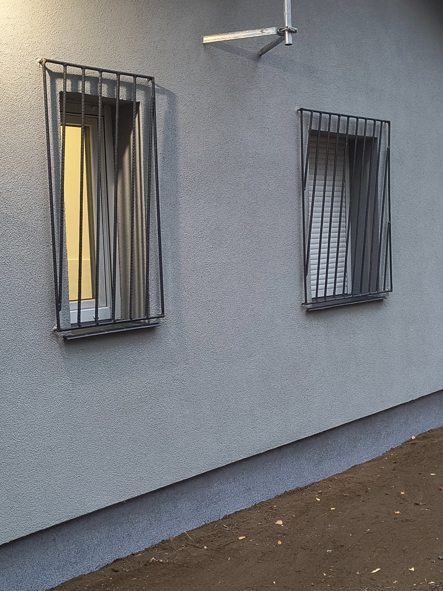 Dwa okna zabezpieczone metalowymi kratami na elewacji budynku z szarym tynkiem strukturalnym, widoczny fragment ziemi przy fundamencie.