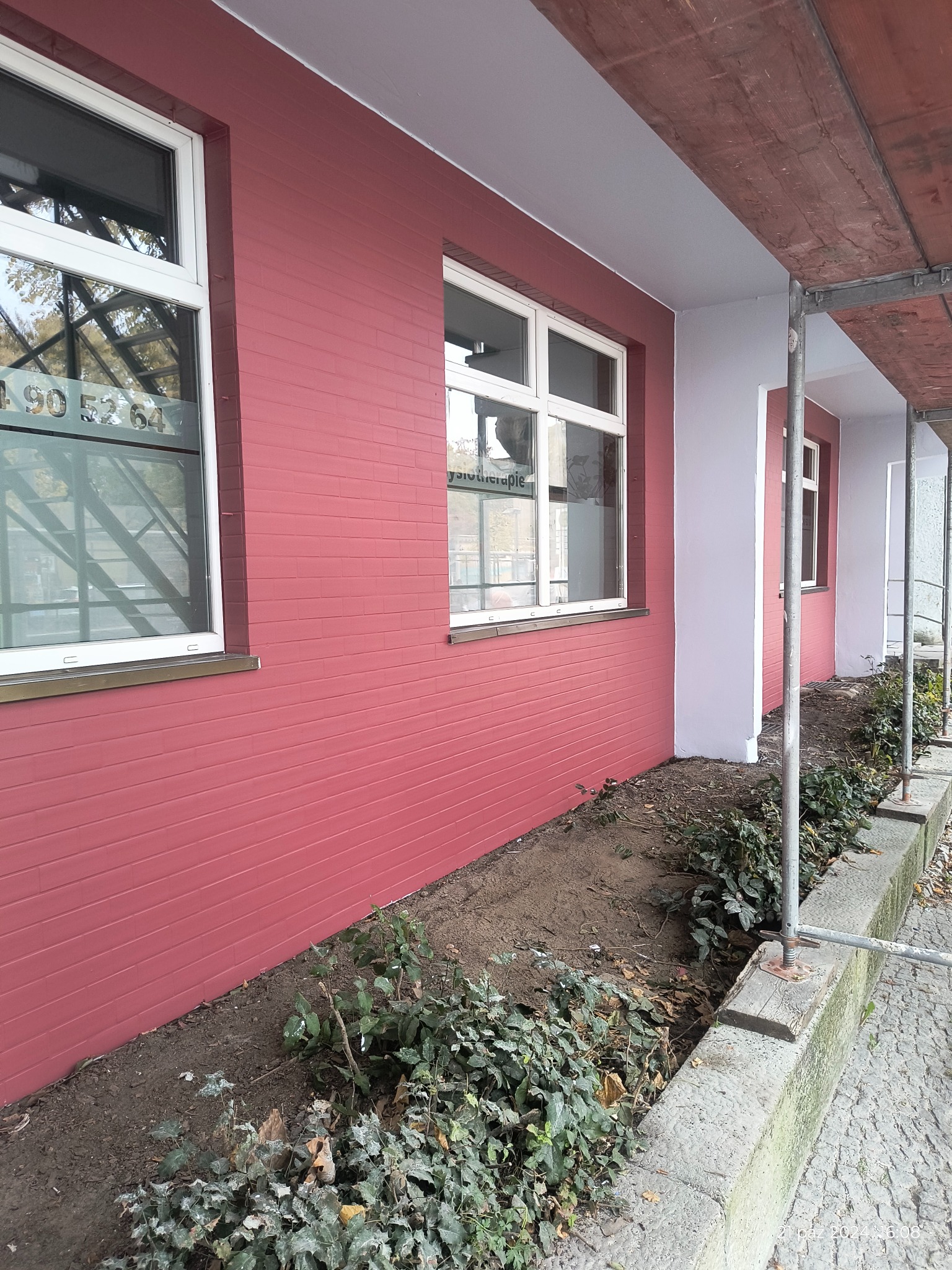 Świeżo pomalowana elewacja budynku w kolorze ceglastym, z białymi ramami okiennymi, widoczne rusztowania i fragment rabaty z zielenią.