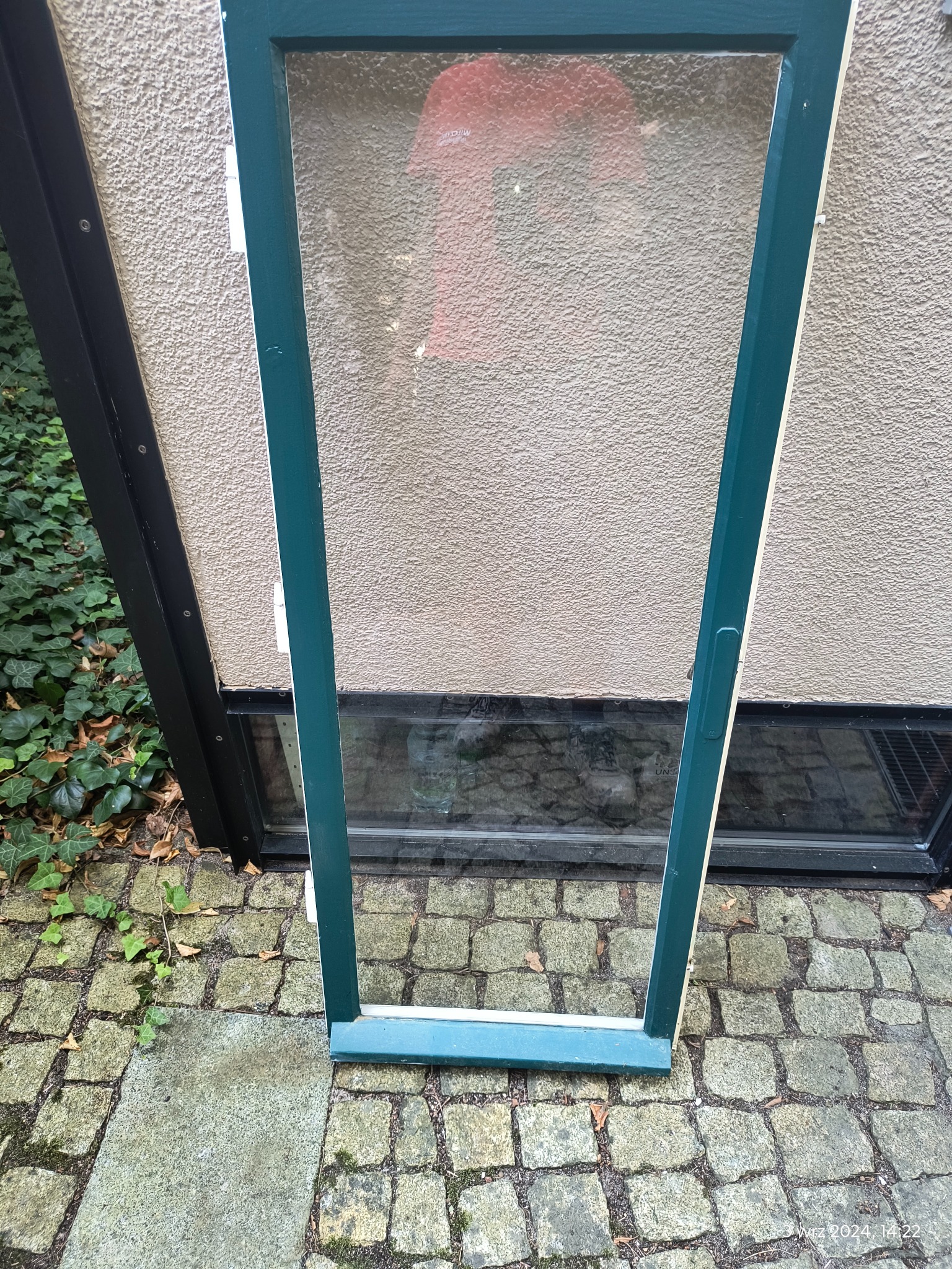 Pomalowana na zielono rama okienna, bez szyby, oparta o ścianę na brukowanym podłożu w Lesznie.
