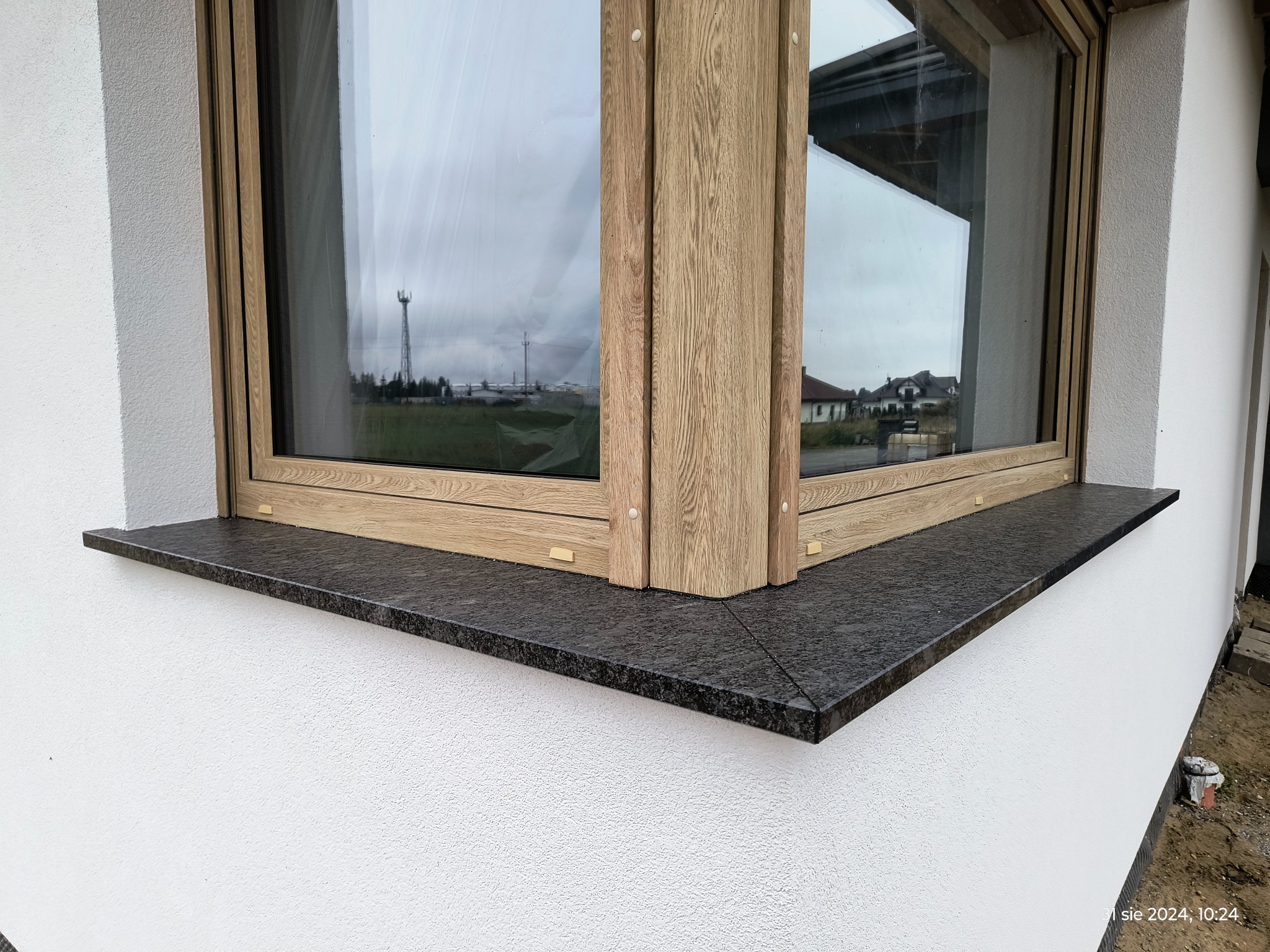 Zamontowany, ciemny, granitowy parapet zewnętrzny pod oknem z drewnopodobną ramą, widok z zewnątrz na białą, teksturowaną elewację budynku.
