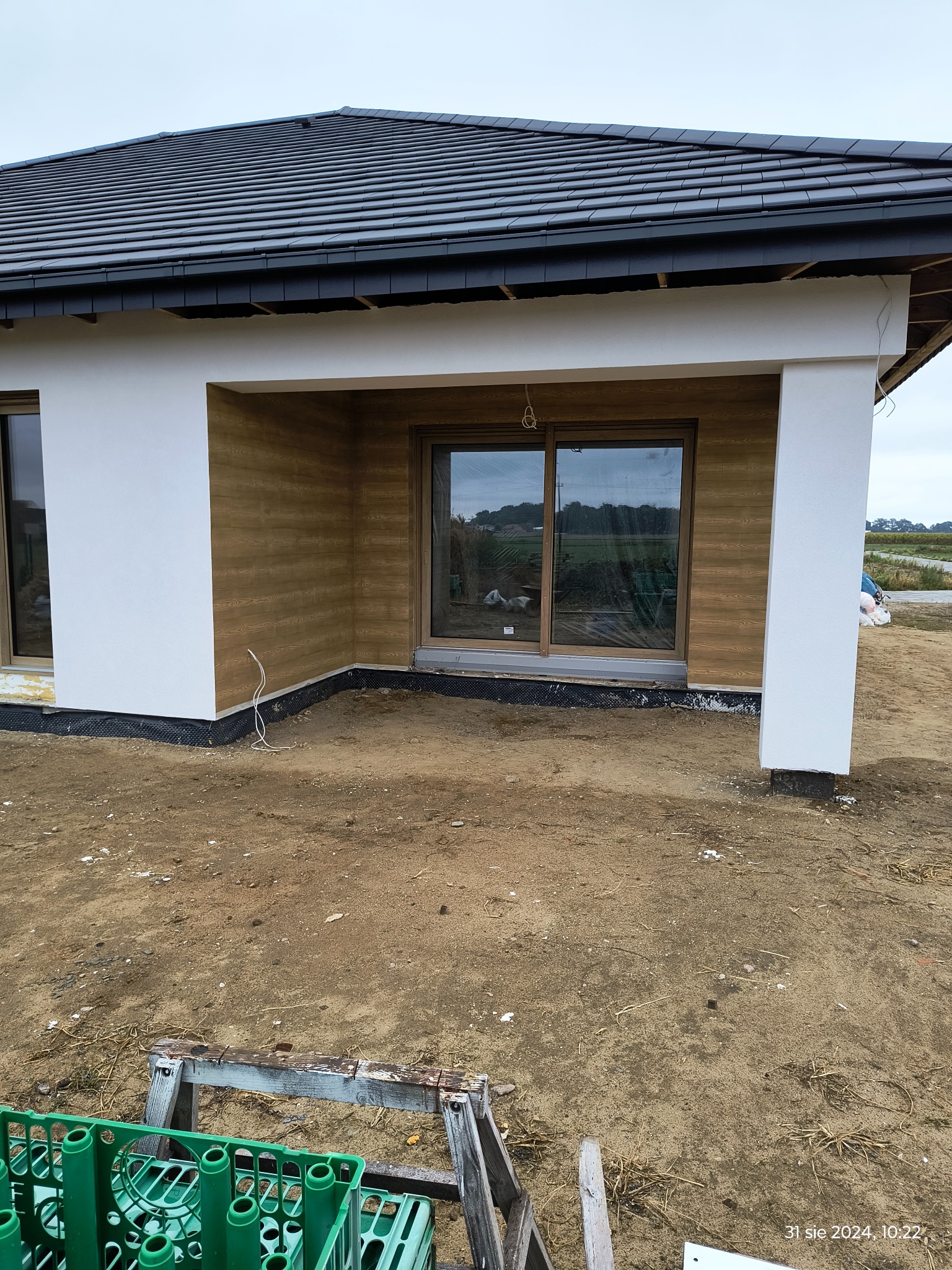 Nowoczesny dom jednorodzinny w trakcie wykończenia elewacji, widoczne okno przesuwne, fragment dachu z ciemnej dachówki i drewnopodobna okładzina elewacyjna.