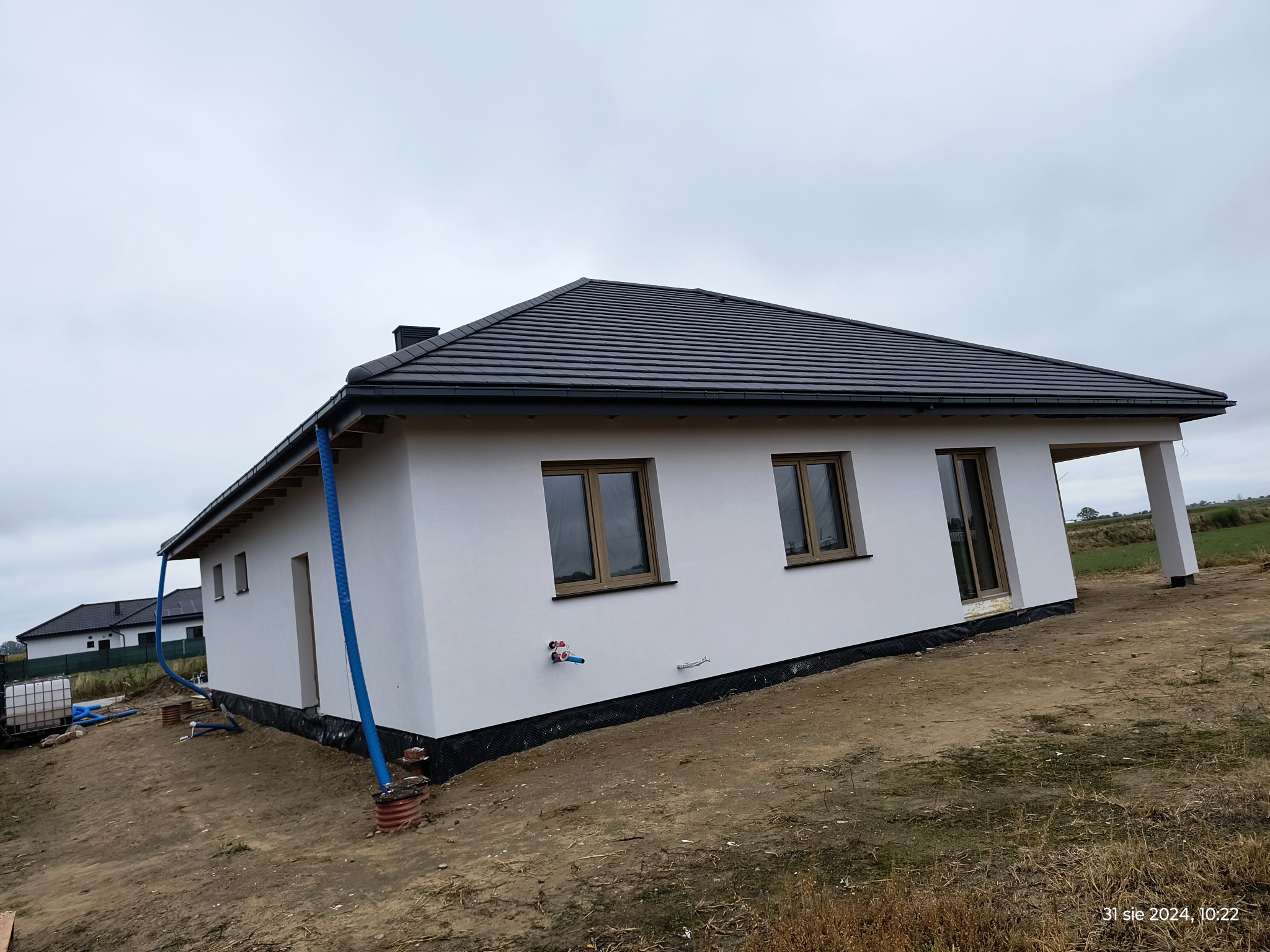 Niedawno otynkowany dom jednorodzinny z grafitową dachówką, widoczne rury spustowe i instalacje na elewacji, teren wokół domu w trakcie zagospodarowywania.
