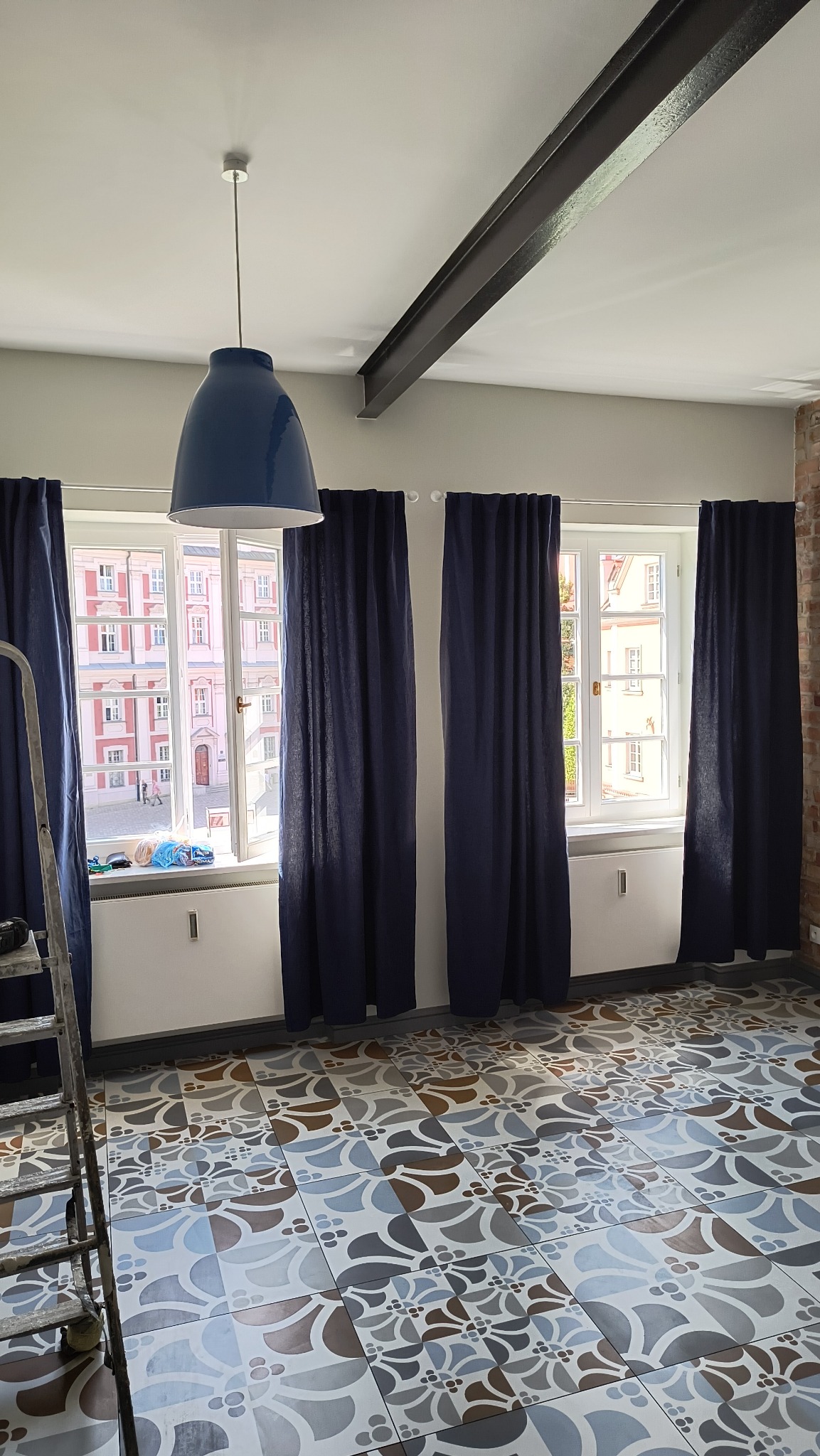 Wnętrze pokoju z wzorzystą podłogą, niebieskimi zasłonami i wiszącą lampą; widoczna drabina malarska i stalowa belka stropowa.