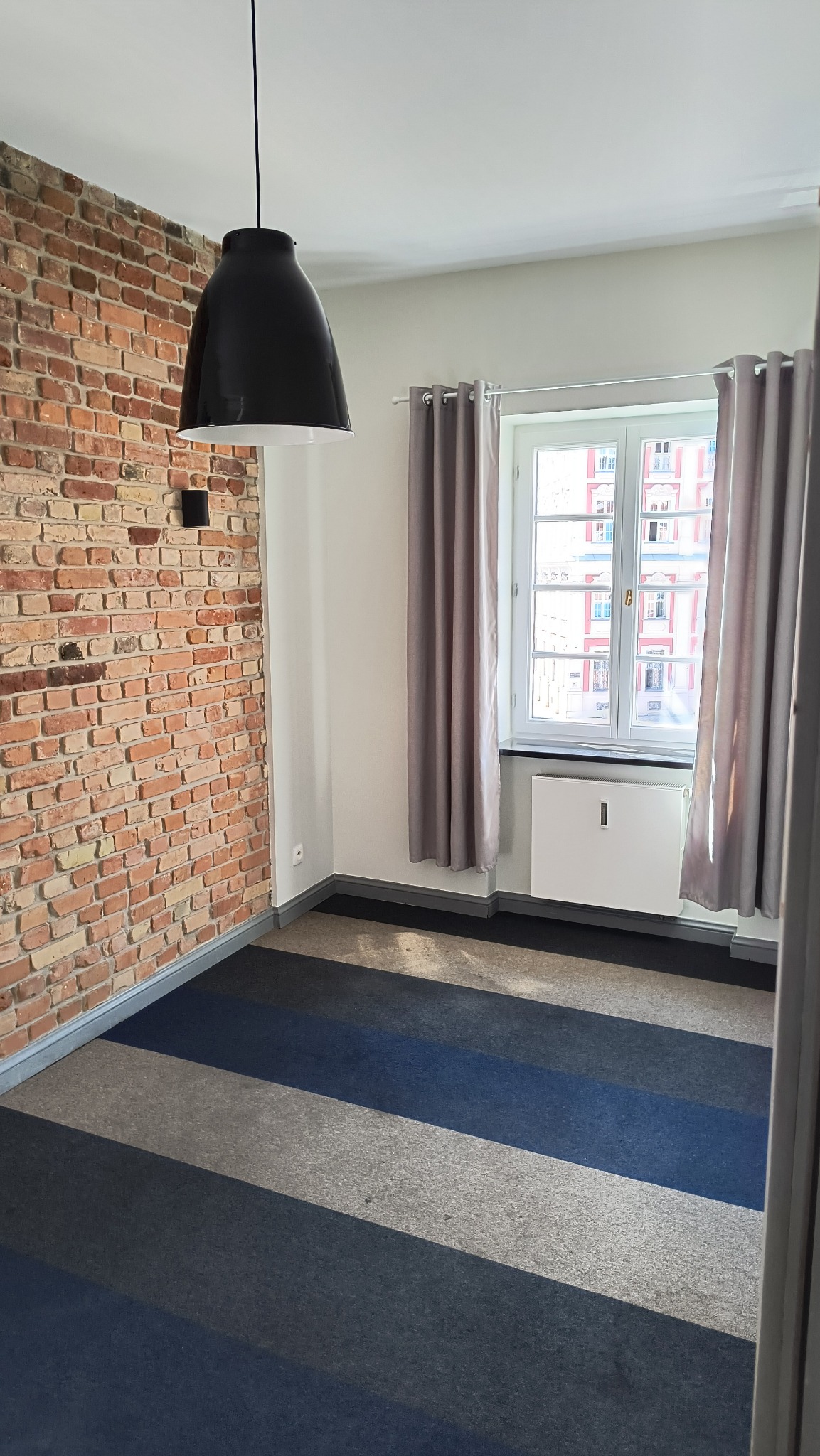 Pusty pokój z odsłoniętą ceglaną ścianą, pasmowym dywanem w odcieniach szarości i niebieskiego, oraz oknem z szarymi zasłonami. Czarna lampa wisząca nad przestrzenią.