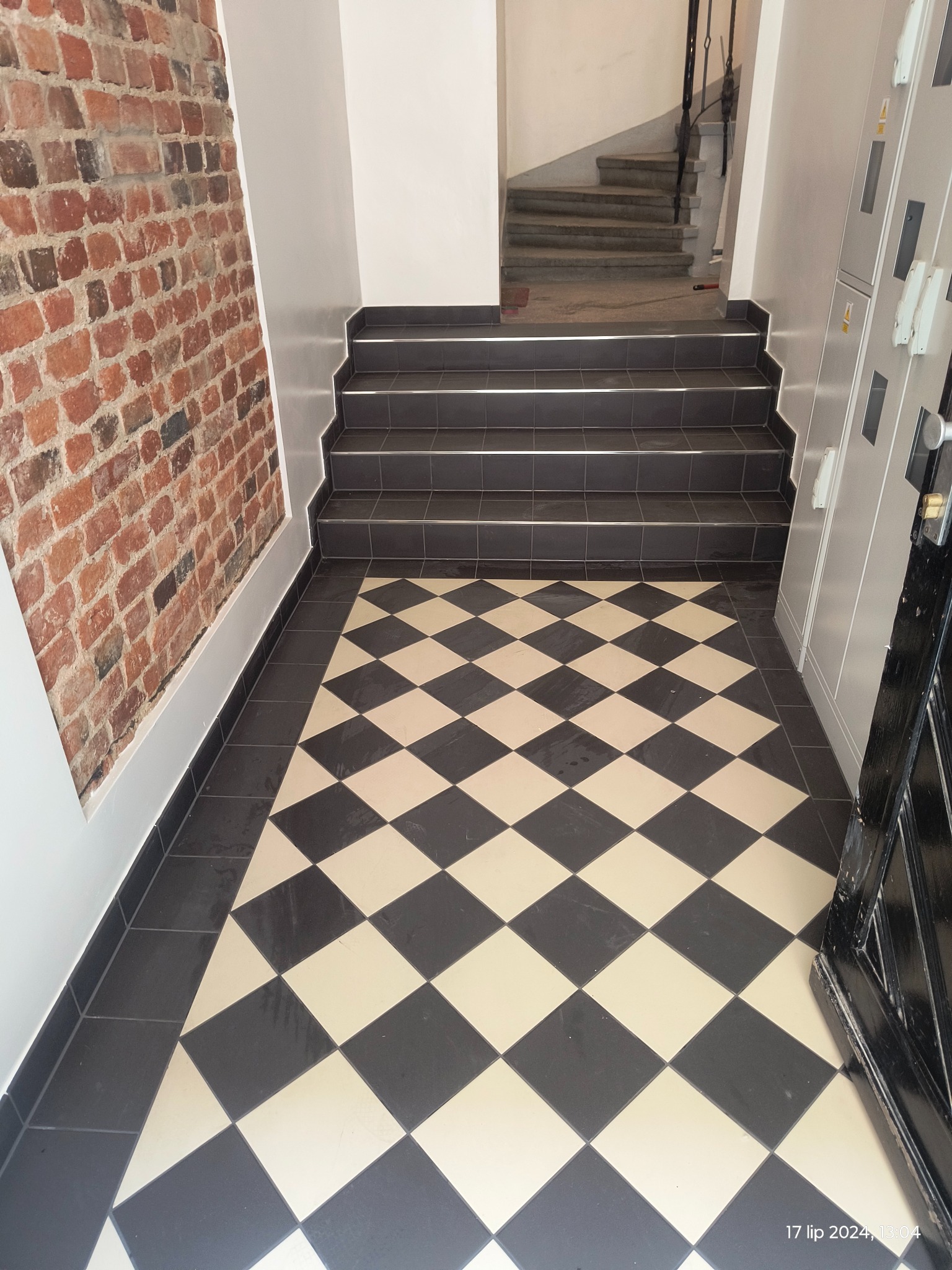 Klatka schodowa z posadzką w czarno-białą szachownicę, widok na schody wykończone ciemnymi płytkami z metalowymi listwami oraz fragment ściany z cegły.