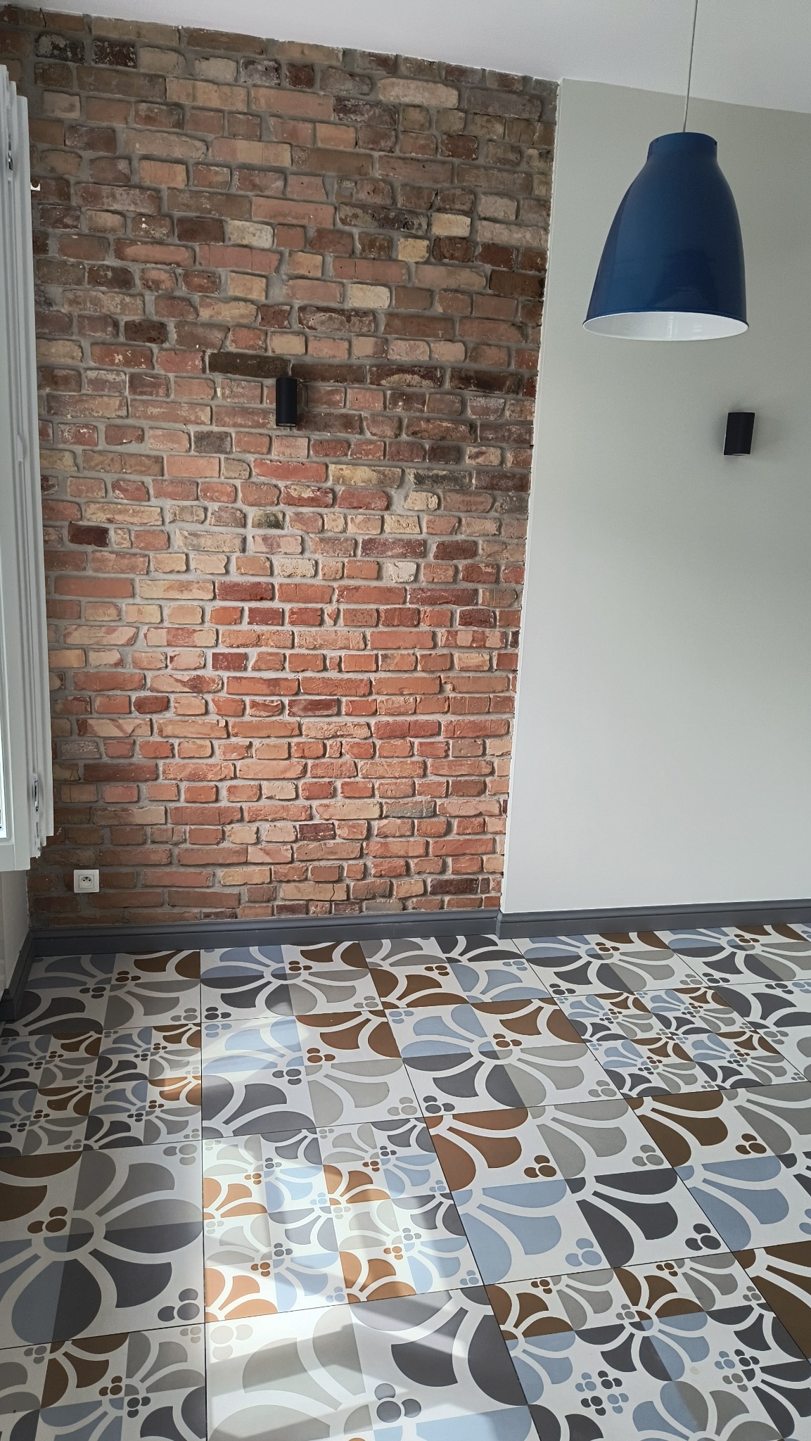 Fragment pokoju z ceglaną ścianą, wzorzystą podłogą i wiszącą niebieską lampą.