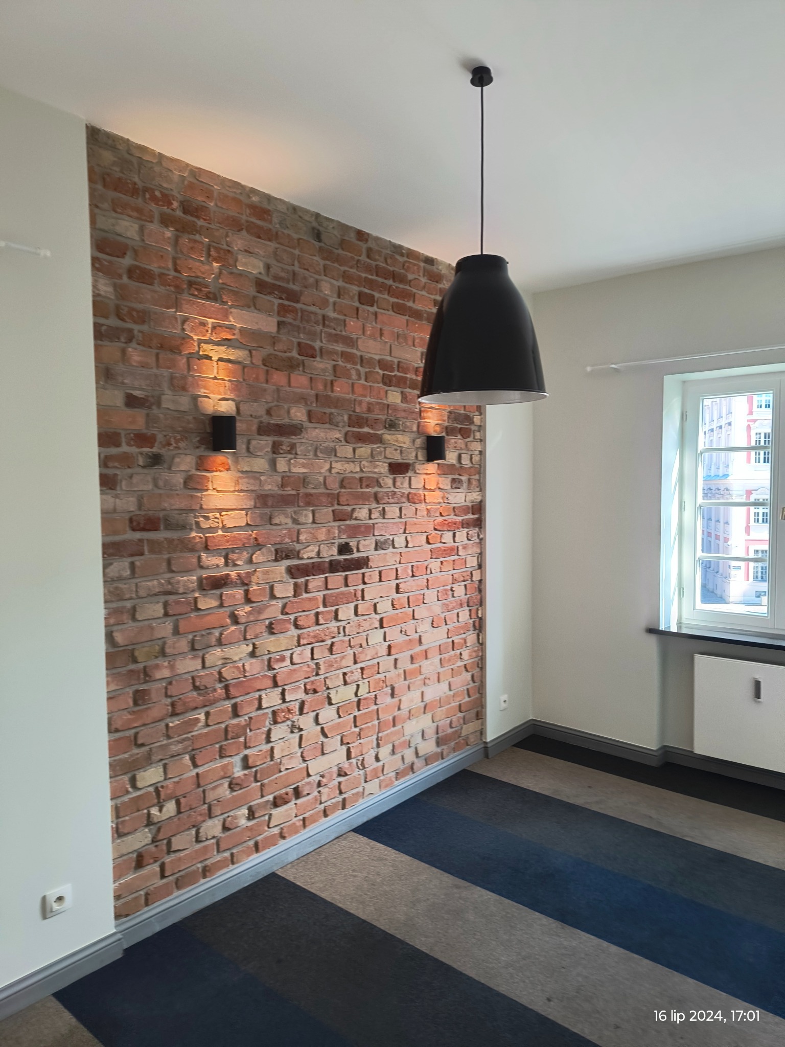 Wnętrze pokoju z dekoracyjną ścianą z czerwonej cegły, oświetloną dwoma kinkietami, z wiszącą czarną lampą i oknem z widokiem na budynek z czerwonej cegły.