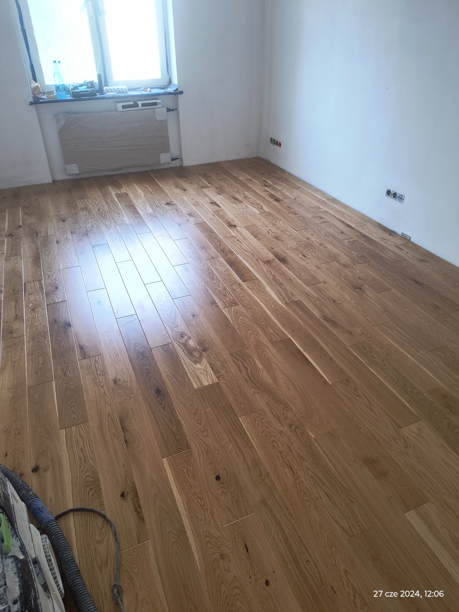 Nowo ułożony parkiet dębowy w jasnym pomieszczeniu z widocznymi gniazdkami elektrycznymi i zakrytym grzejnikiem, częściowo widoczna maszyna do obróbki parkietu.