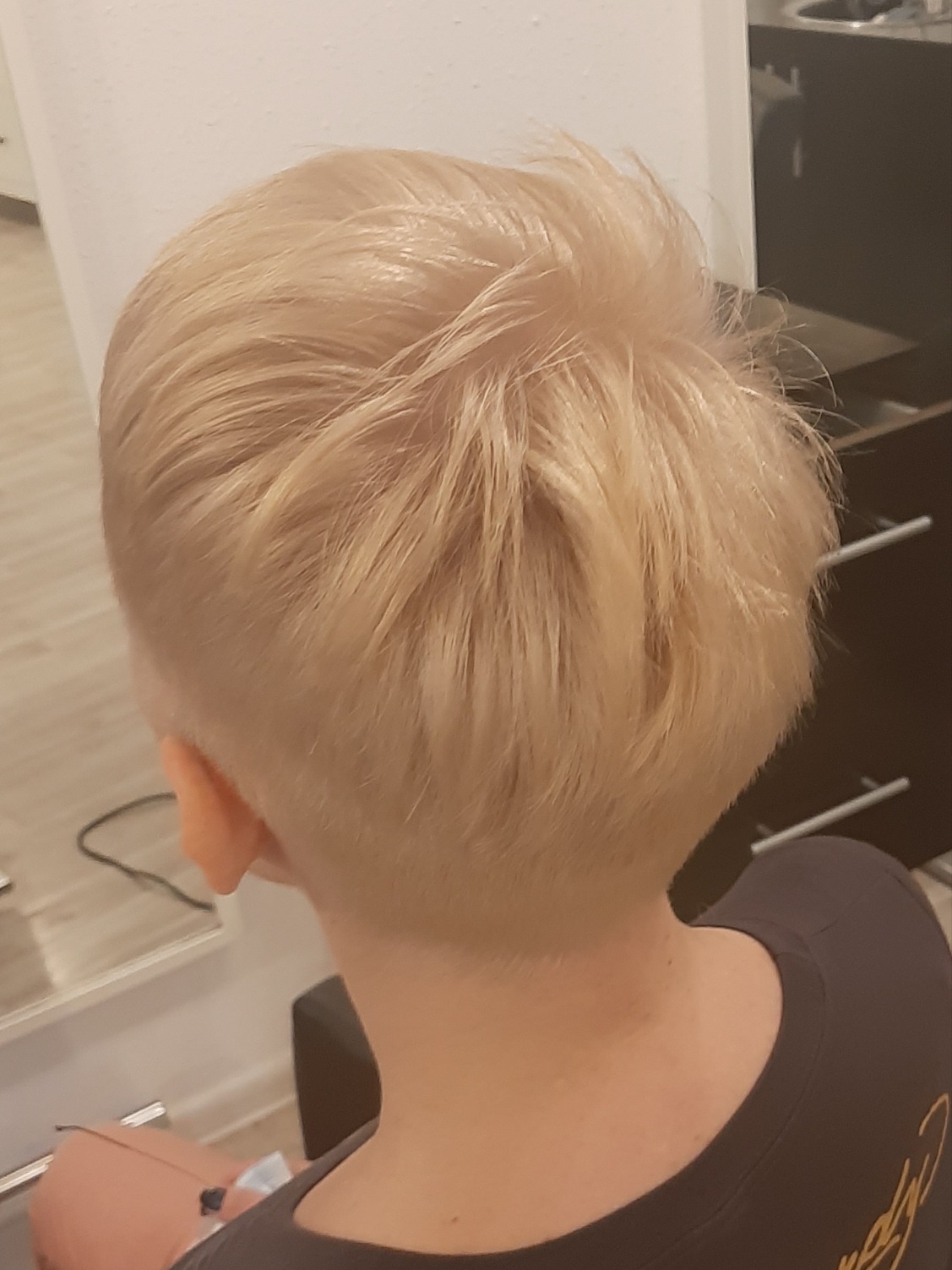 Blond włosy po strzyżeniu pixie cut, widoczne z tyłu głowy, z krótkimi bokami i dłuższą górą, delikatne cieniowanie.